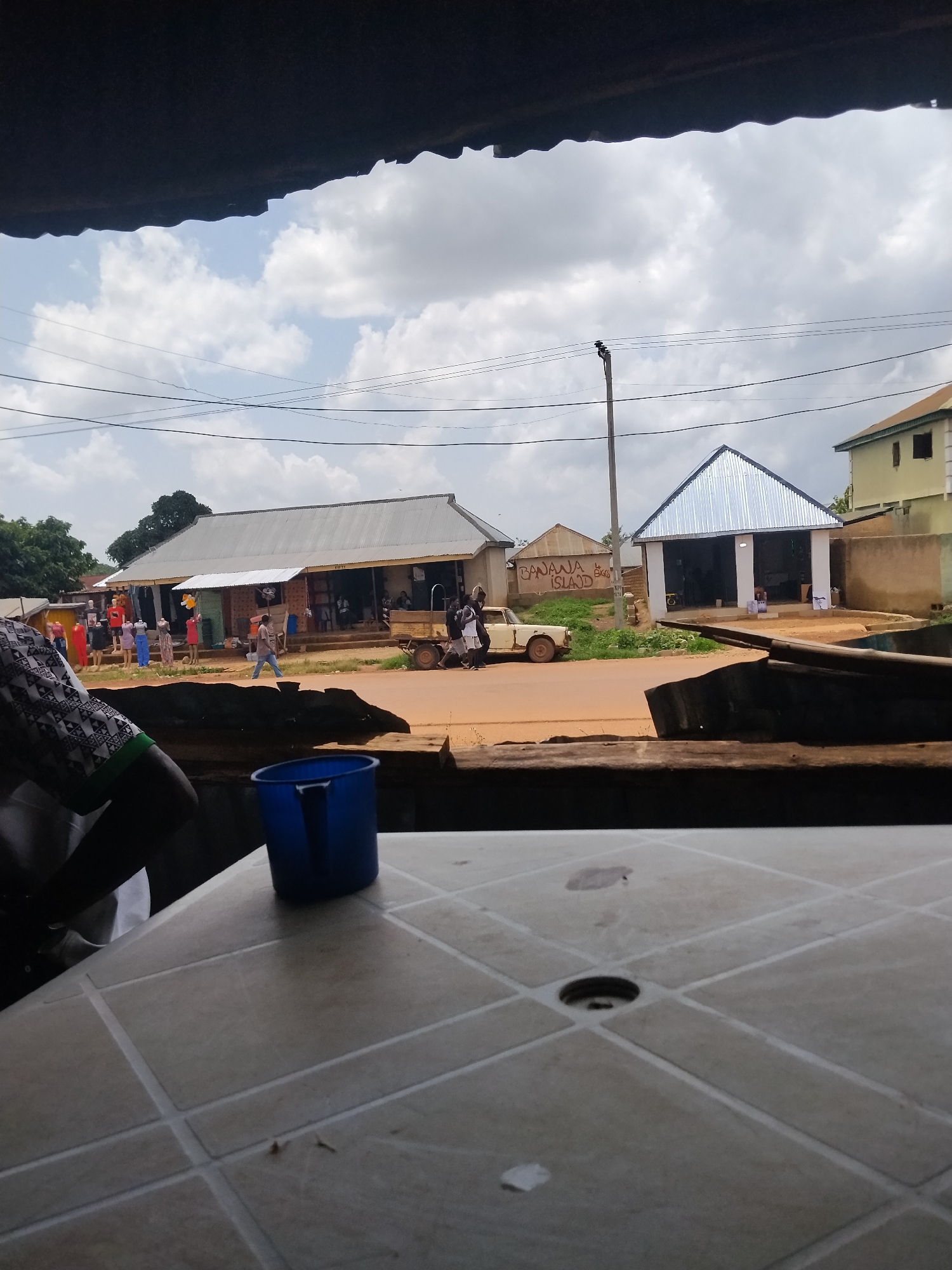The image depicts a street scene in Durumi, Nigeria. In the foreground, a portion of a light-colored, tiled table is visible, with a blue plastic mug placed on it. The middle ground shows a street with a few vehicles and people. To the left, several mannequins are displayed outside a shop, showcasing various clothing items. Further down the street, a white car is parked. Several buildings are present, including one with a silver corrugated metal roof and another with a light green facade. Power lines crisscross the sky. The background reveals more buildings and trees under a cloudy sky. The overall impression is that of a modest commercial area in a Nigerian town.