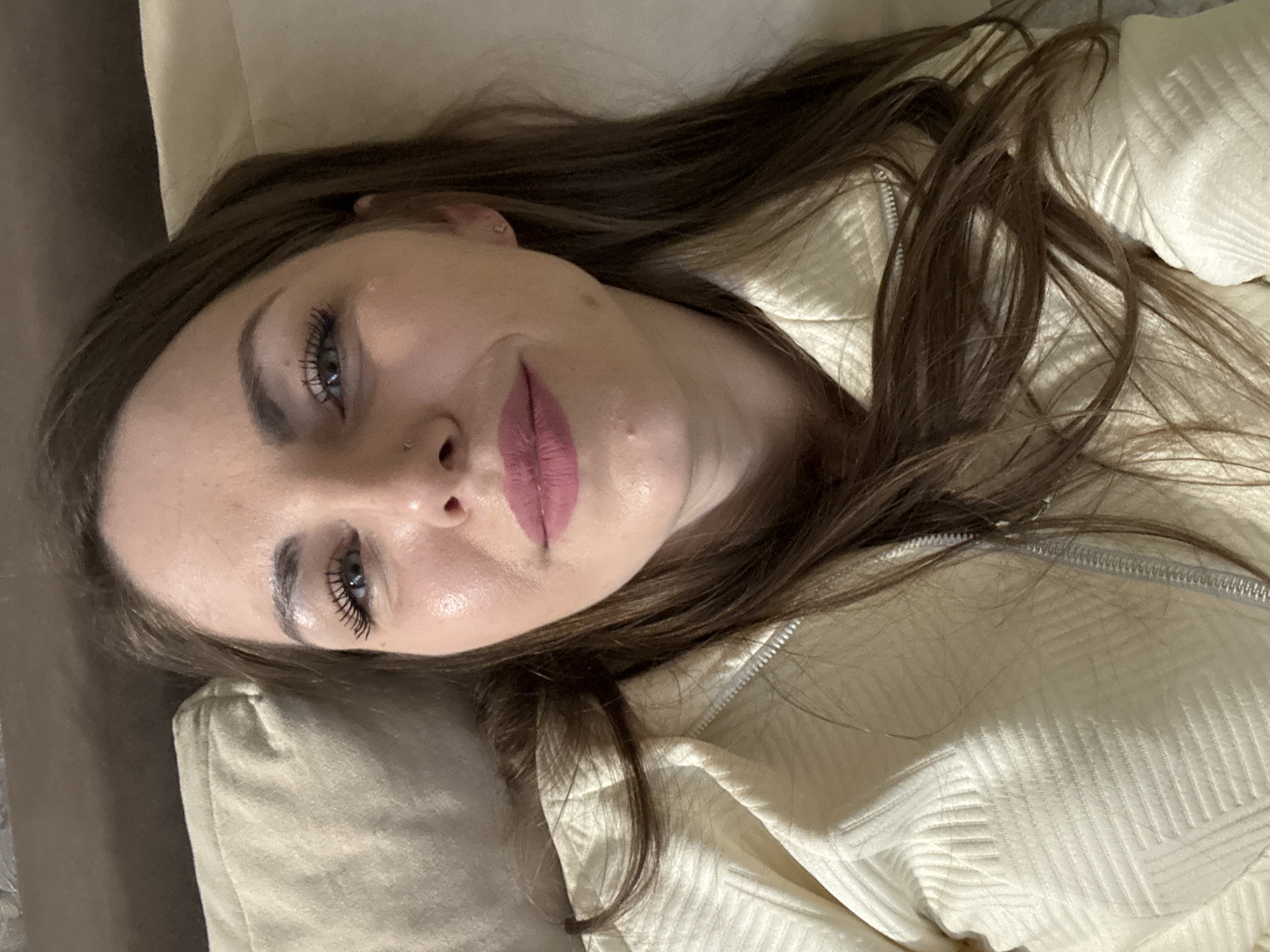 A young woman with brown hair is lying down, facing the camera. She has light skin, gray eyes, and is wearing lipstick. Her head rests on a beige surface, possibly a cushion or part of a sofa. She is wearing a white zippered jacket. The location is not discernable from the image.