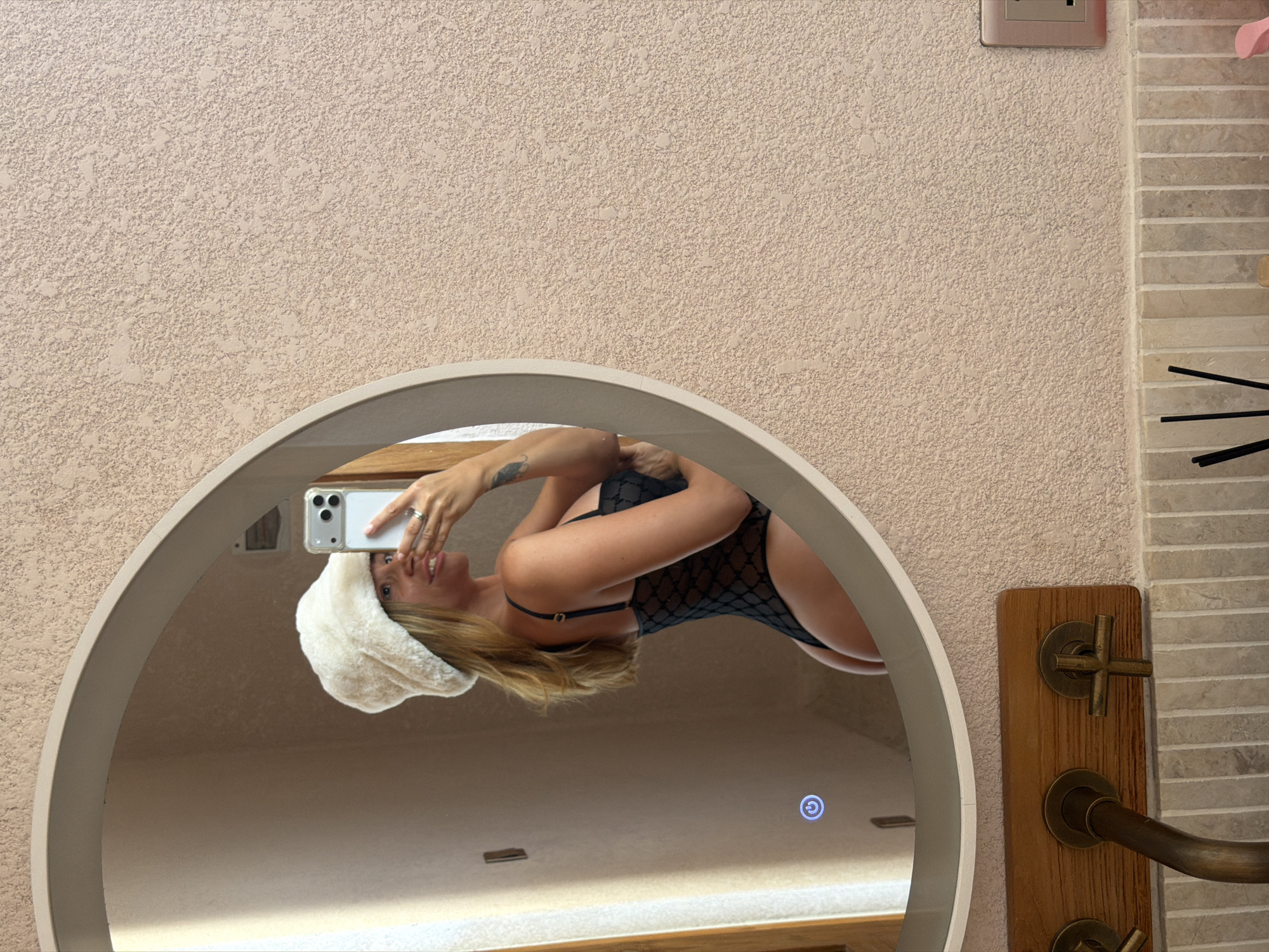 A woman in a white furry hat and black lingerie is taking a mirror selfie in a bathroom. The mirror is oval-shaped and framed by a light-colored border. The woman's left side is facing the camera, and she is holding a smartphone in her right hand. She is smiling and looking at her reflection. The background of the bathroom is tiled with a textured, light-colored material. There is a wooden panel with faucets visible below the mirror. The image was taken in a bathroom in the city of Mengwi, Indonesia.