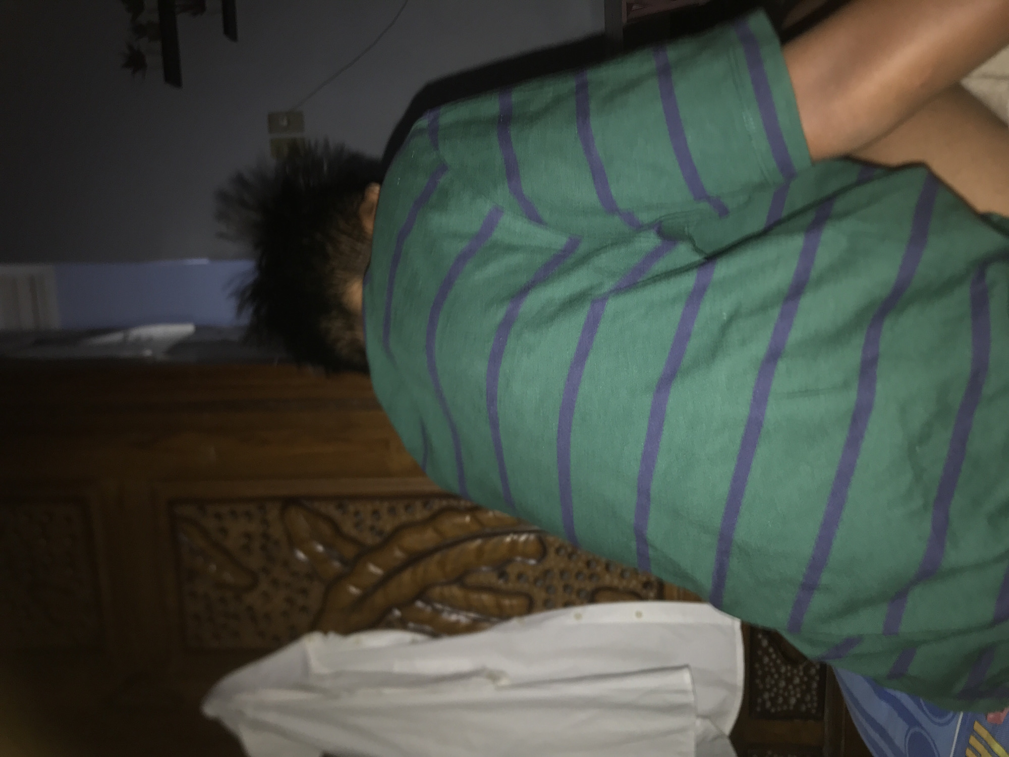 The image shows a person lying face down on a bed in a dimly lit room. The person is wearing a green and blue striped t-shirt and their head is turned to the left. The bed has a wooden headboard with a decorative carved pattern. A white shirt lies on the bed near the person. The specific location in Lhokseumawe, Indonesia is not discernible from the image.