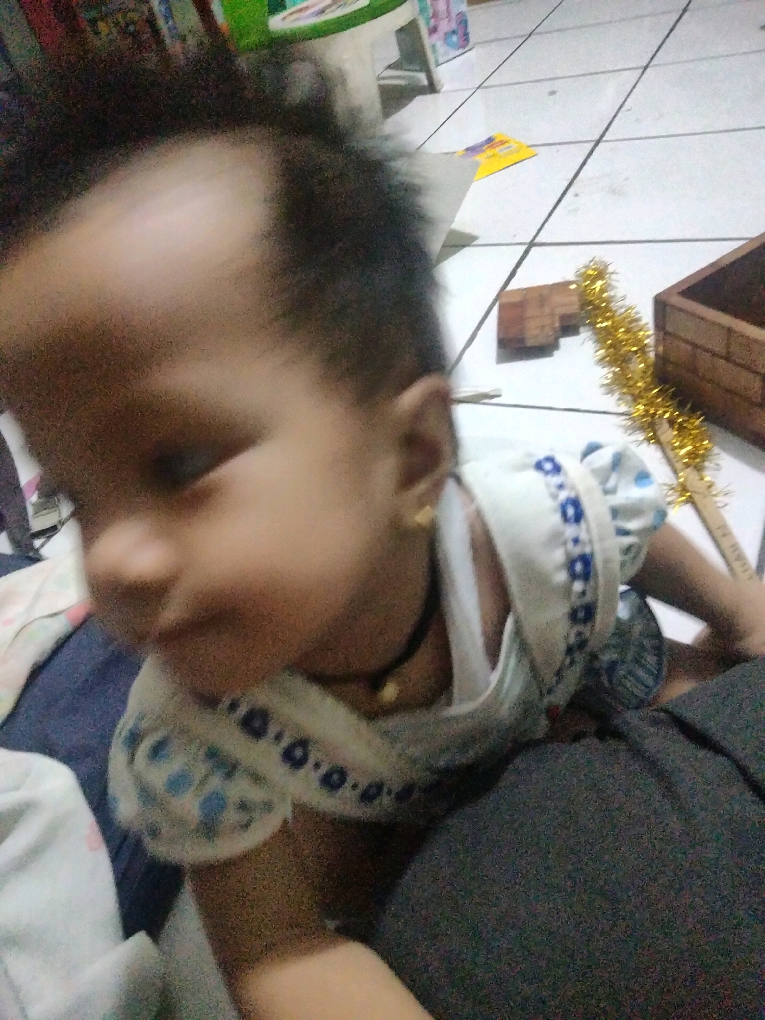 In the city of Suka Sari, Indonesia, an infant is captured in a close-up, low-angle shot, lying on a tiled floor. The infant's face is partially obscured, with a notable bald patch on the forehead and dark, wispy hair around the sides. They are wearing a white and blue patterned garment, a black choker necklace, and a small gold earring in their left ear. The background reveals a tiled floor with visible grout lines, and scattered objects including a wooden structure, a yellow pamphlet, and what appears to be gold tinsel. A large dark surface, possibly clothing or bedding, occupies the foreground. The lighting is dim, and the image exhibits a grainy quality.