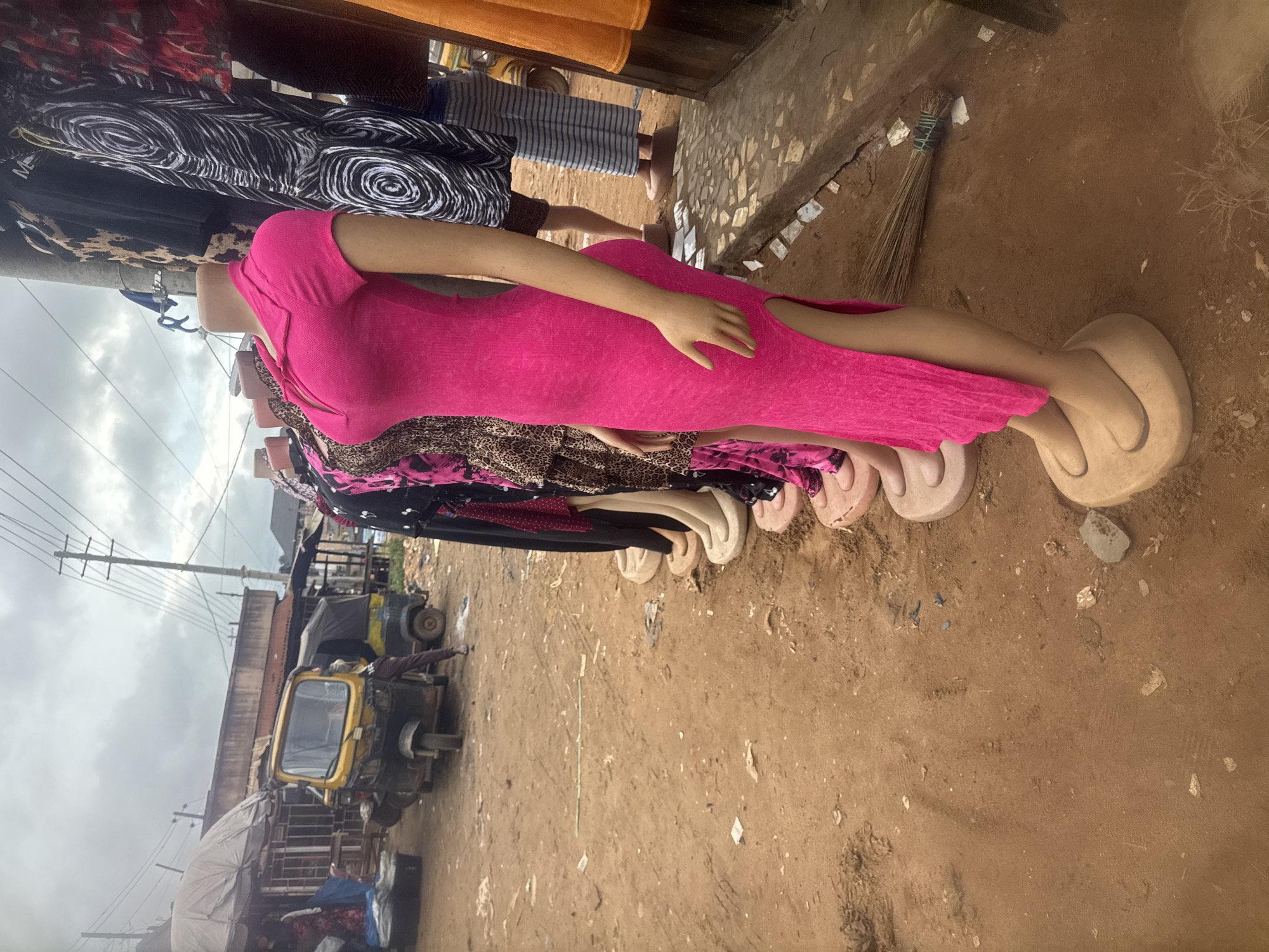 The image depicts a street market scene in Nkpor, Nigeria, with a focus on mannequins displaying clothing. In the foreground, several mannequins are lined up, showcasing various dresses, including a bright pink one with a slit up the side. The mannequins are made of a flesh-toned material and stand on circular beige bases. Behind the mannequins, clothing items are hung on display racks, featuring patterns like animal print and abstract designs.
In the mid-ground, a yellow and black auto-rickshaw, commonly known as a Keke Napep, is parked on the dirt-covered ground. A person wearing dark pants and a light-colored shirt is seen near the auto-rickshaw, appearing to interact with it. Further back, other stalls or shops can be seen with awnings and roofing structures. The environment is dusty and appears to be an outdoor marketplace. The sky is overcast. To the right of the mannequins, a broom made of natural fibers tied with green string lies on the ground.