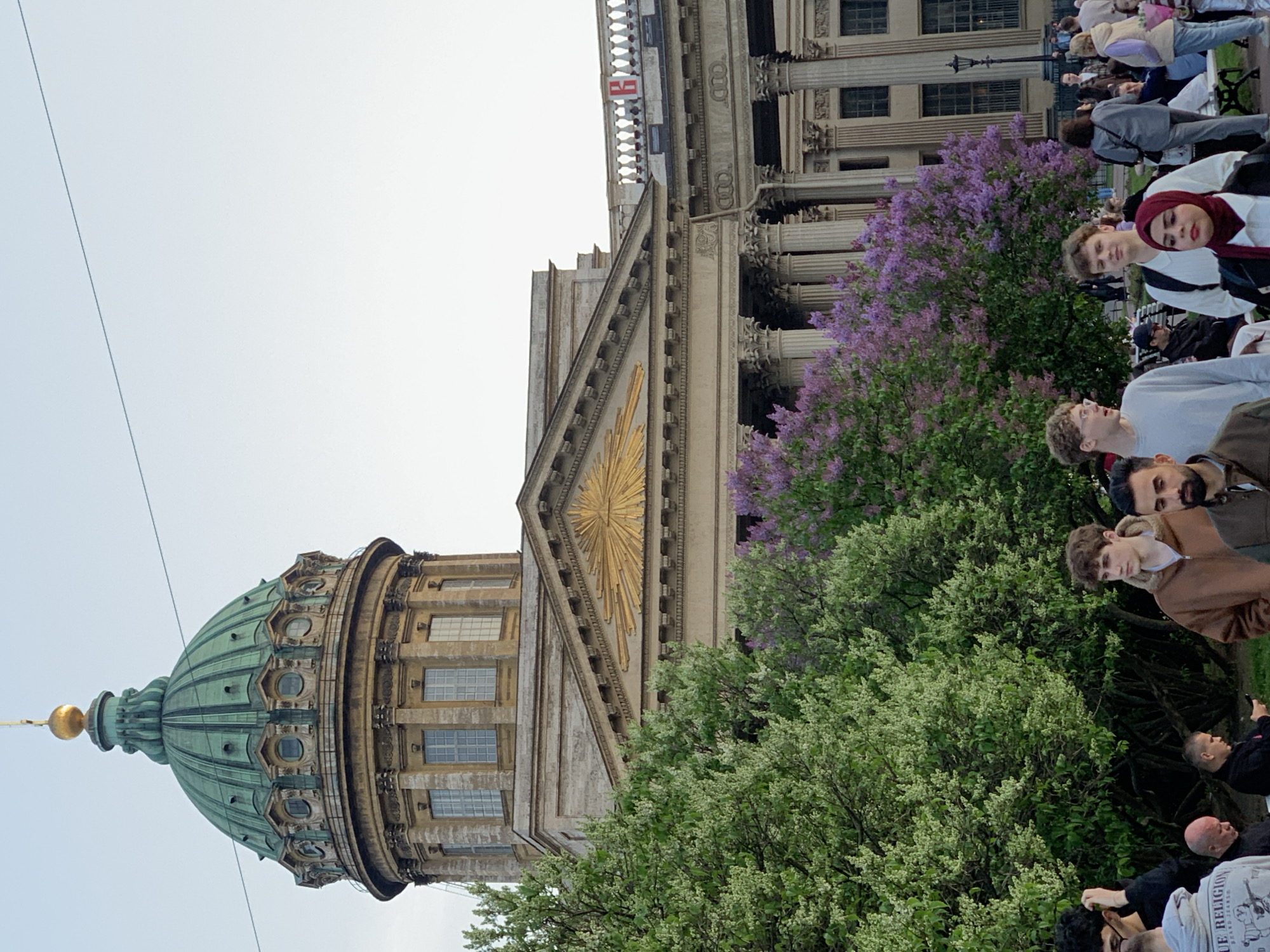 The image depicts the Kazan Cathedral in Saint Petersburg, Russia. The cathedral's dome is visible on the left side of the frame, with the main facade and columns extending towards the right. A golden emblem is positioned prominently on the front of the building. Several people are standing in front of the cathedral, amidst blooming trees and bushes. They appear to be tourists or visitors, observing the structure. The sky is overcast.