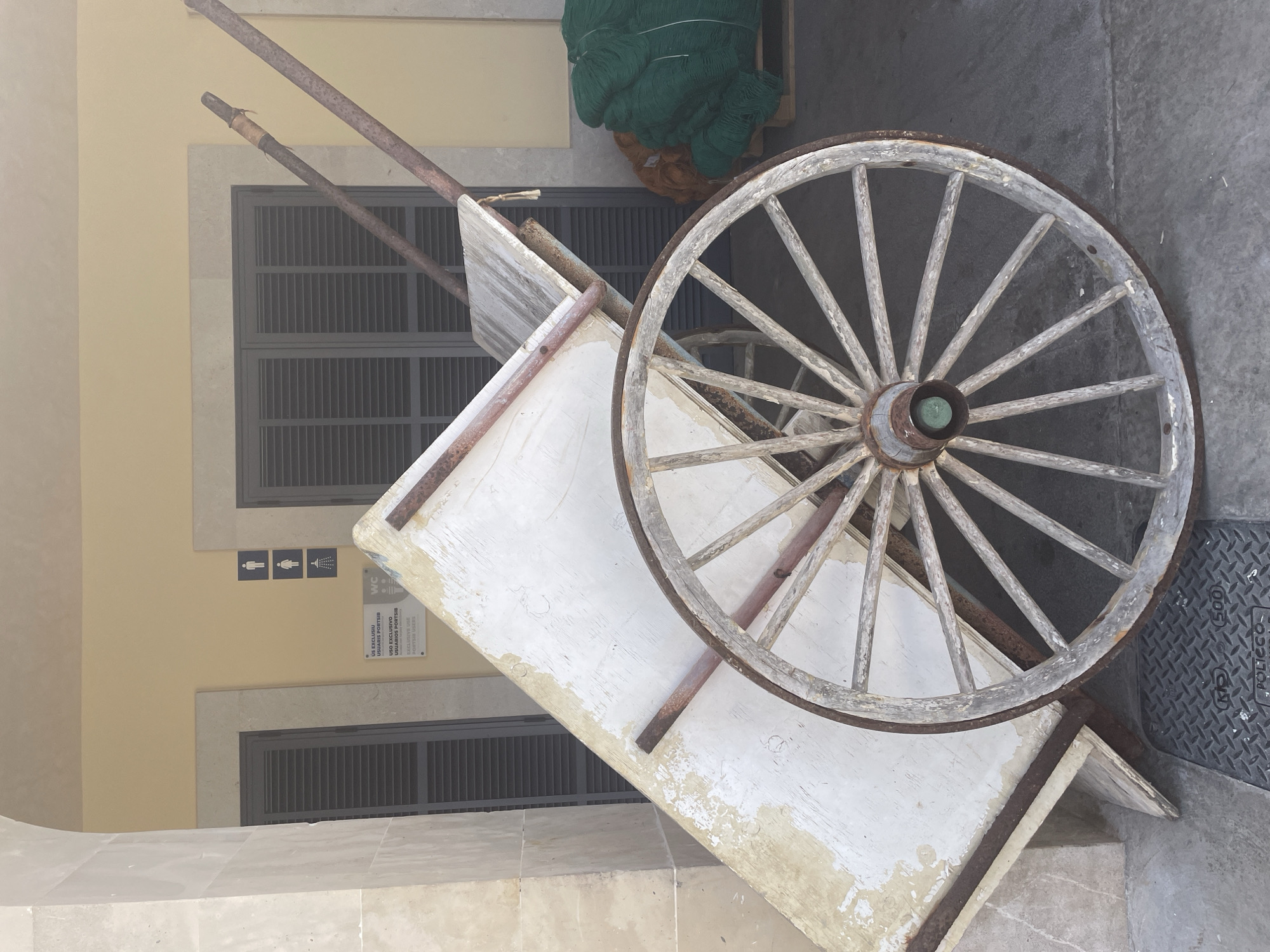 The image captures a rustic, weathered wooden cart with a single spoked wheel. The cart's body, painted white with visible wear and tear, is tilted upwards, resting against a building with large, louvered shutters. Two metal poles extend from the cart, suggesting it was used for transport.
In the background, a pale yellow building features a sign with men's and women's restroom icons, along with a shower icon, indicating a public facility. Below the icons, a bilingual sign (Spanish and Catalan) reads "US EXCLUSIU USUARIS PORTSÍB / USO EXCLUSIVO USUARIOS PORTSÍB", signifying exclusive use for port users. Further to the right, stacks of green and orange netting are visible on a pallet, possibly related to fishing activities.
The scene appears to be set in an outdoor area with a concrete floor. The overall impression is one of a functional, perhaps historic, piece of equipment in a seaside or port location. Based on the Spanish signage, the location is likely Cala Figuera, Spain, a small fishing village in Mallorca.