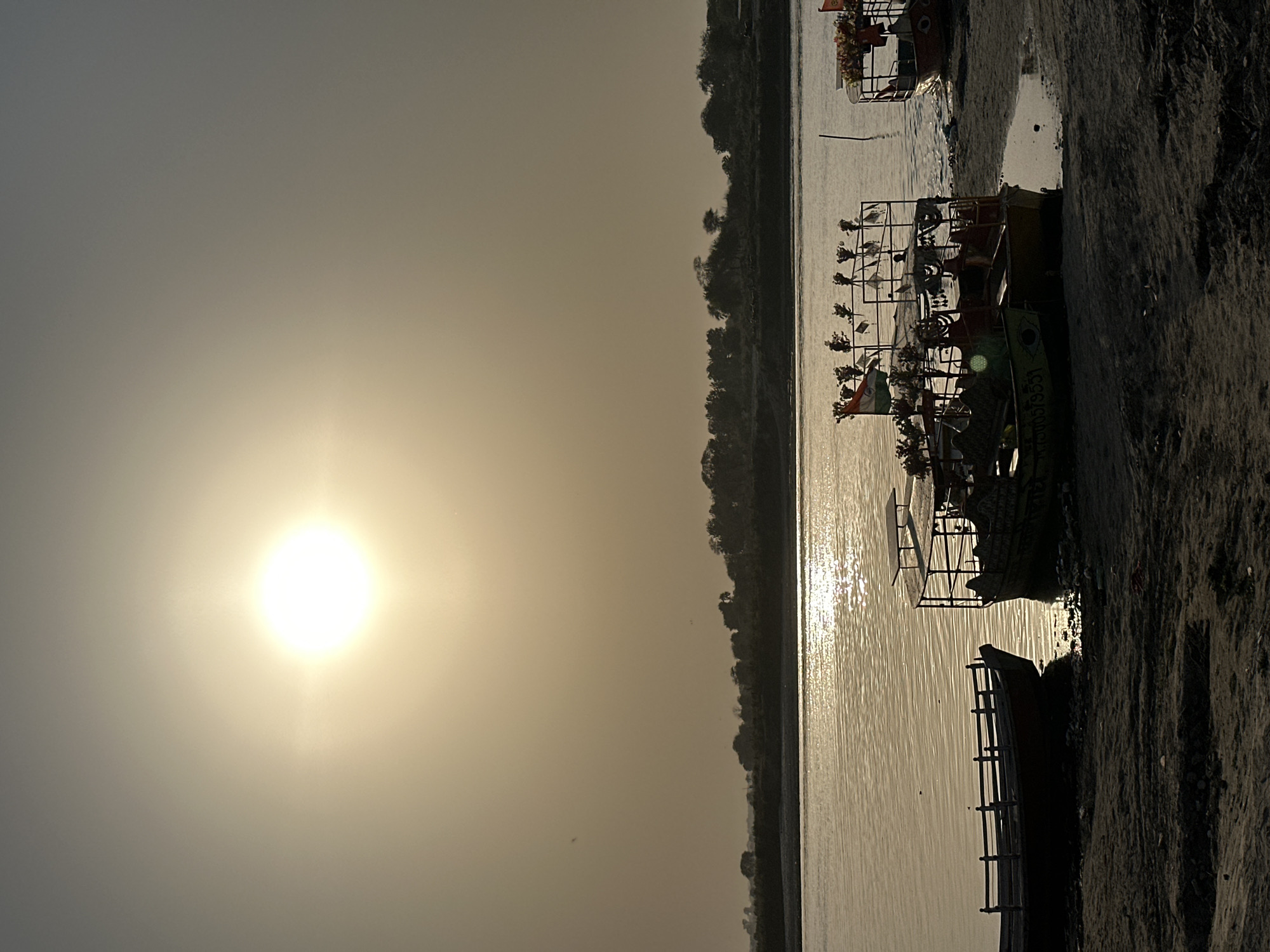 The photograph captures a tranquil scene at Vrindavan, India. The sun dominates the sky on the left side of the image, creating a golden glow over the landscape. A river stretches horizontally across the frame, reflecting the sunlight. Along the riverbank on the right, several boats are docked. One boat features an Indian flag among decorative elements, adding a cultural touch to the scene. The riverbank itself is textured and contrasts with the smooth water surface. The image presents a peaceful, sunlit view of the riverfront in Vrindavan.
