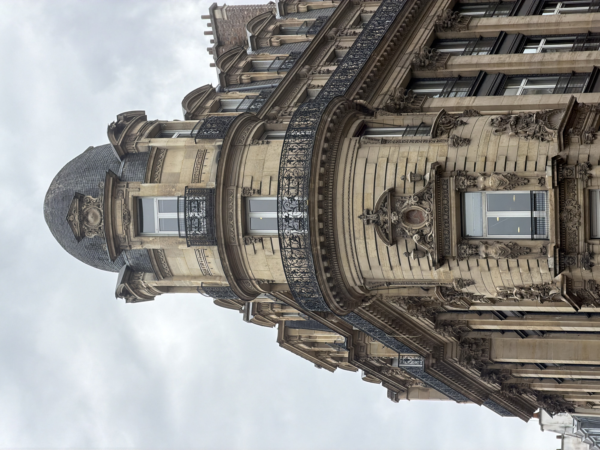 The image depicts a low-angle view of a building in Paris, France. The architecture is ornate, featuring light-colored stone, decorative carvings, and black wrought-iron balconies. The building has a rounded corner capped with a dome covered in grey shingles. The sky is overcast. No people are visible.