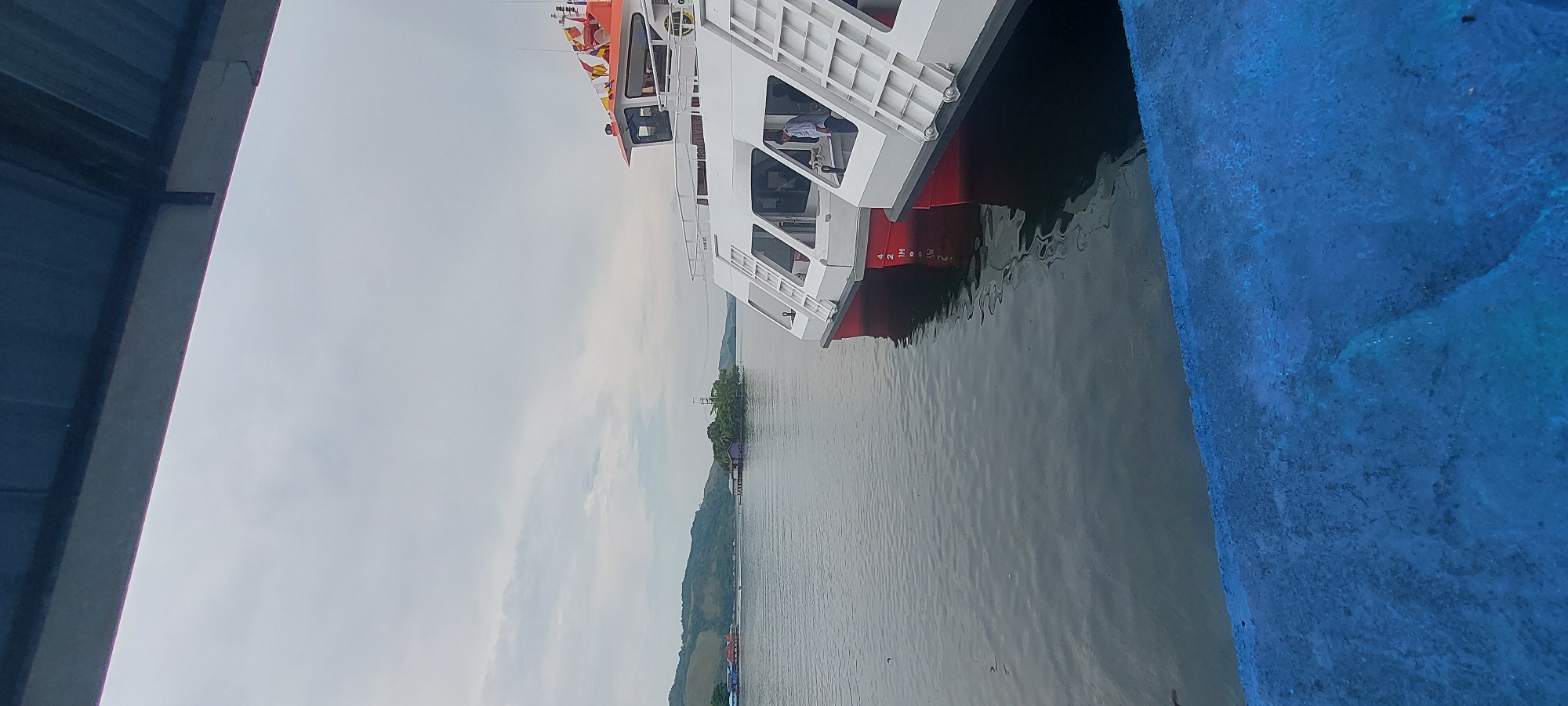 This image depicts a serene waterscape in Asei Besar, Indonesia, likely at a dock. In the foreground, a blue railing is visible, suggesting a vantage point from a pier or elevated walkway. To the right, the stern of a white passenger ferry with red trim is partially visible, indicating it is docked. The ferry has multiple windows and staircases leading to upper decks. The water is calm and reflects the overcast sky. In the middle ground, a small, tree-covered island with a few structures is situated in the lake. Further in the background, rolling green hills form the shoreline, with more buildings and structures visible along the coast. The sky is cloudy, contributing to a subdued natural light.