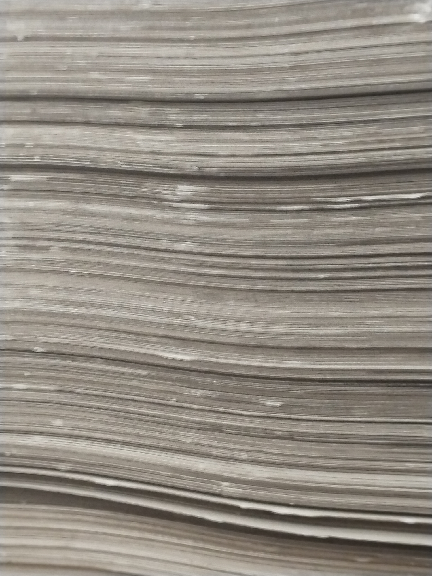 A close-up, abstract shot shows a stack of paper or thin, flat materials layered horizontally. The texture is uniform with subtle variations in shade, suggesting a dull, neutral color like grey or beige. The lighting is even, casting no discernible shadows, which flattens the image and emphasizes the repetitive, linear pattern of the stacked items. The overall impression is one of density and order. The image does not contain any elements that would suggest a specific location within the city of Гожскі сельскі Савет, Belarus, or any human presence.