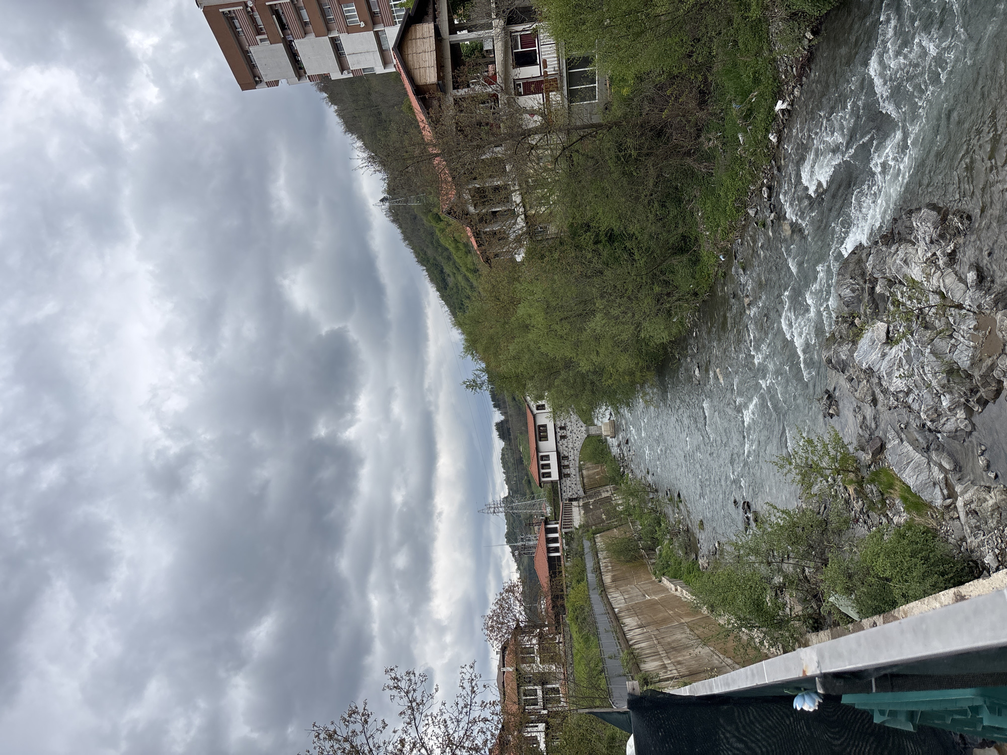 The image captures a river valley scene in Zlatograd, Bulgaria, under an overcast sky. A river with turbulent, white-capped water flows from the right side of the frame towards the left, through a natural landscape.
On the left bank of the river, a concrete embankment or pathway is visible. Beyond this, several buildings with traditional red-tiled roofs and light-colored facades are nestled among green trees and bushes. One larger, multi-story building with brown and white sections is partially visible towards the upper left, suggesting a more urban presence. The buildings extend up the green, tree-covered hillsides that form the backdrop.
The right bank of the river consists of rocky terrain and dense green foliage. In the immediate foreground, a grey metal railing suggests the photograph was taken from a bridge or elevated viewpoint overlooking the river.
The sky is completely covered by thick, grey clouds, indicating an overcast day. A tall metal power transmission pylon stands on a distant hillside, hinting at infrastructure within the natural setting. No people or discernible text are visible in the image. The overall impression is one of a peaceful, somewhat dramatic natural landscape influenced by human settlement.