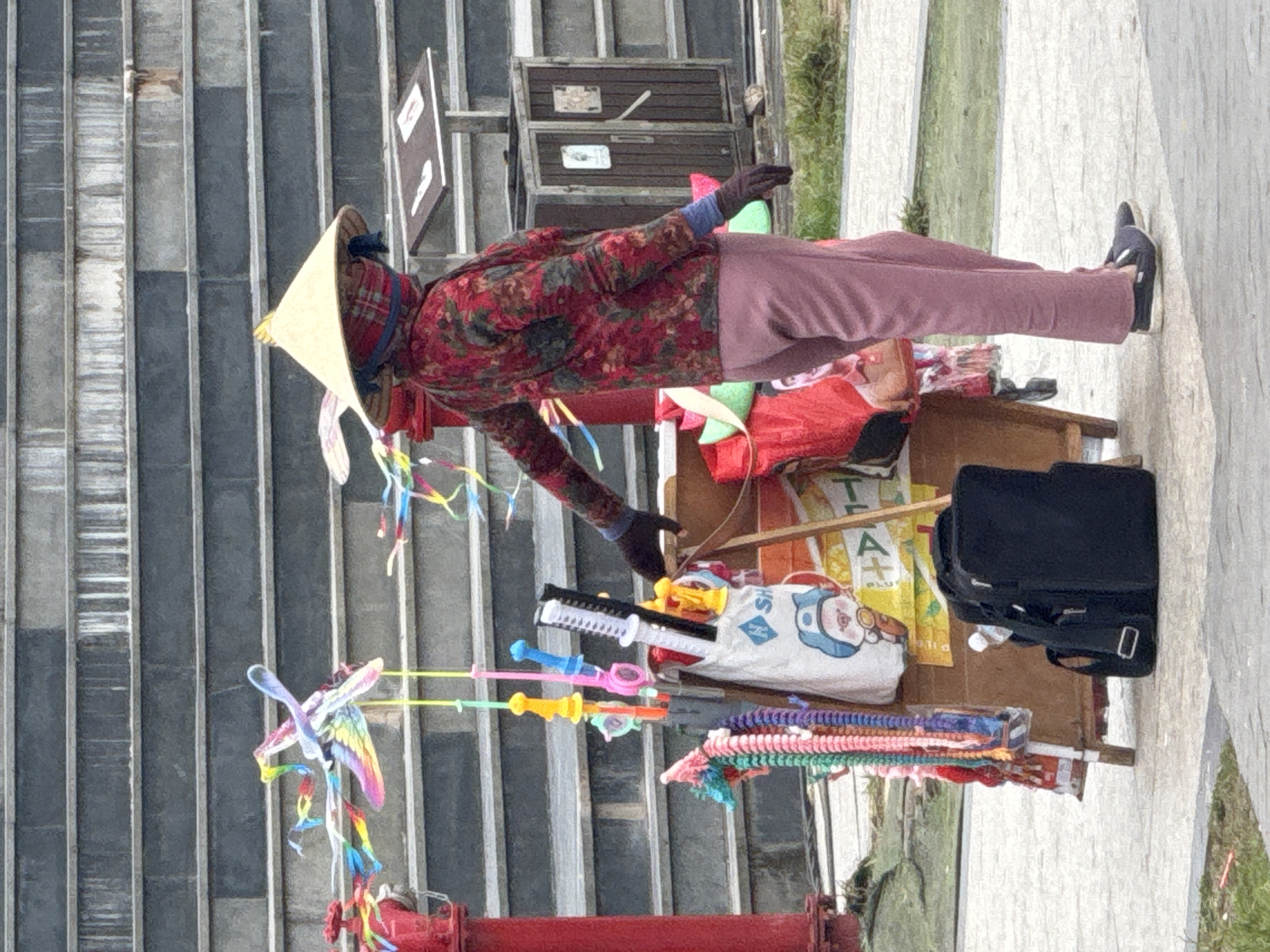 A person wearing a conical hat and a red patterned long-sleeved shirt is standing next to a cart filled with toys and accessories. The cart displays colorful items such as kites, beaded necklaces, and toy swords. A black backpack is placed on the ground next to the cart. In the background, a set of stone steps and a trash can are visible. The scene appears to be in an outdoor urban environment, possibly a public space in Da Lat, Vietnam, given the conical hat and the style of the cart and merchandise. The ground is paved with concrete slabs.