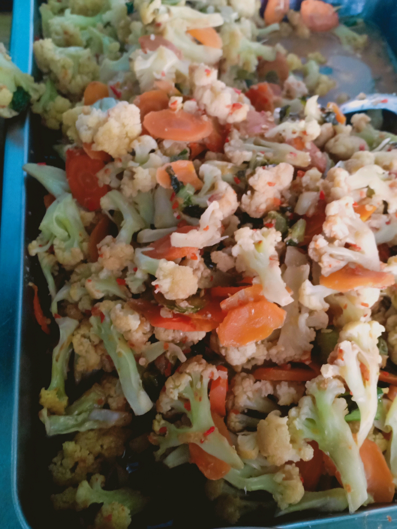 A close-up, high-angle shot shows a metal tray filled with a colorful mixture of stir-fried vegetables, primarily cauliflower florets and sliced carrots. The vegetables appear to be seasoned with small red chili flakes, visible sprinkled throughout the dish. The cauliflower is light yellow and white, while the carrots are a vibrant orange. The stems of the cauliflower are a pale green. The tray itself is metallic and appears to be part of a serving setup. This dish is likely being prepared or served in Candipuro, Indonesia, given the context.