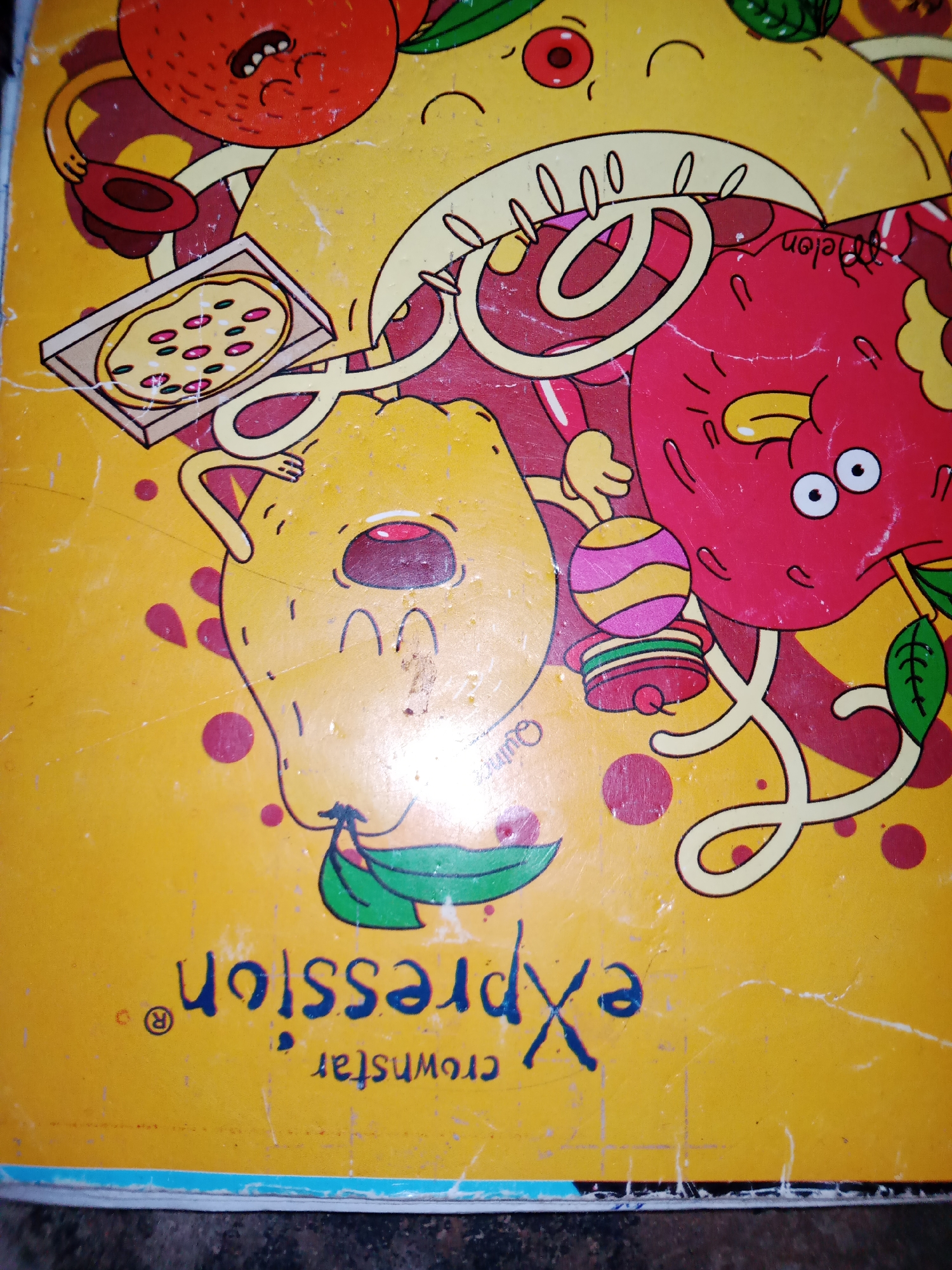 The image is a close-up, overhead shot of a notebook cover with a cartoonish, vibrant design. The background is a bright yellow, with scattered pink polka dots. The main subjects are anthropomorphic fruits or food items, rendered in a playful, exaggerated style with bold outlines.
On the left, a distressed-looking orange character is shown, holding a slice of pizza in a cardboard box. To its right, a large, round yellow character, possibly a lemon or a melon, with wide eyes and an open mouth, is depicted. Further to the right, a red, apple-like character with wide eyes and a determined expression is present. These characters are intertwined with red, looping lines, suggesting movement or connection.
There is a small, striped ball on a stack of colorful rings near the red character. The overall scene evokes a sense of fun and perhaps a celebration or a lively gathering. The emotions conveyed by the characters range from distress (orange) to joy or excitement (red character).
Visible text on the cover includes "expression" in large, blue, stylized lettering, written upside down. Below this, also upside down, is "crownstar" and a registered trademark symbol. Another small, faint text appears to be "Quincy" near the yellow character.
The image appears to be taken indoors, possibly on a desk or table, given the slightly worn and scratched surface of the notebook cover and the dark background visible at the bottom edge. The bright colors and playful illustration suggest it might be a children's notebook or a promotional item. The location context of Jauro Yinu, Nigeria, does not provide any direct visual cues within the image itself.