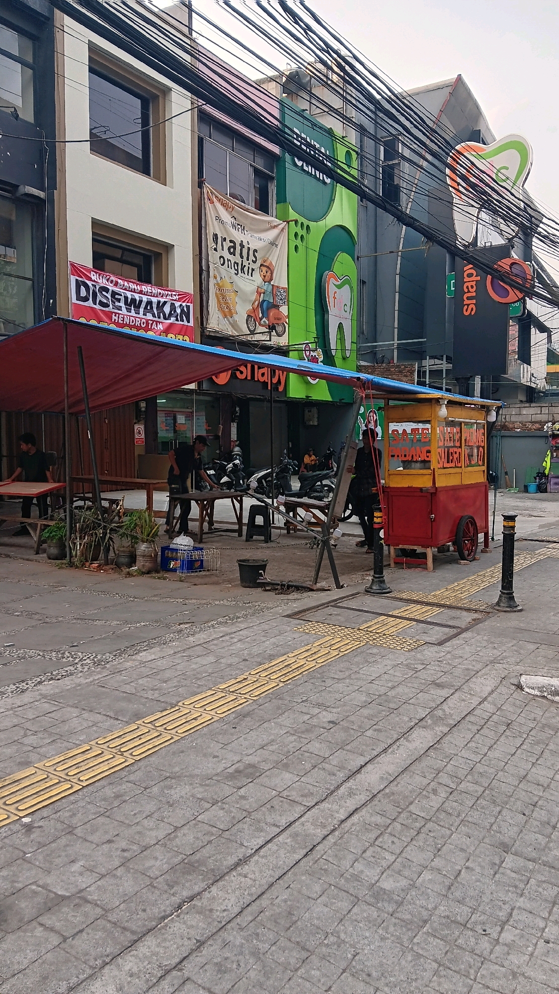 The image depicts a street scene in South Jakarta, Indonesia, possibly on a sidewalk in front of commercial establishments. In the foreground, a red and yellow food cart with the words "Sate Padang Seler KJo" is visible, indicating it sells Sate Padang, a type of Indonesian satay. Behind the cart, a red awning covers a seating area with tables and chairs, where several people appear to be present, some standing and some seated. Motorcycles are parked in the background. The buildings lining the street feature signage, including one for "DENTAL CLINIC" and another with the word "snapv". Overhead, a dense network of electrical wires crisscrosses the sky. The sidewalk is paved with gray bricks and features yellow tactile paving for visually impaired pedestrians. The overall impression is of a busy urban environment with street food vendors and commercial activity.