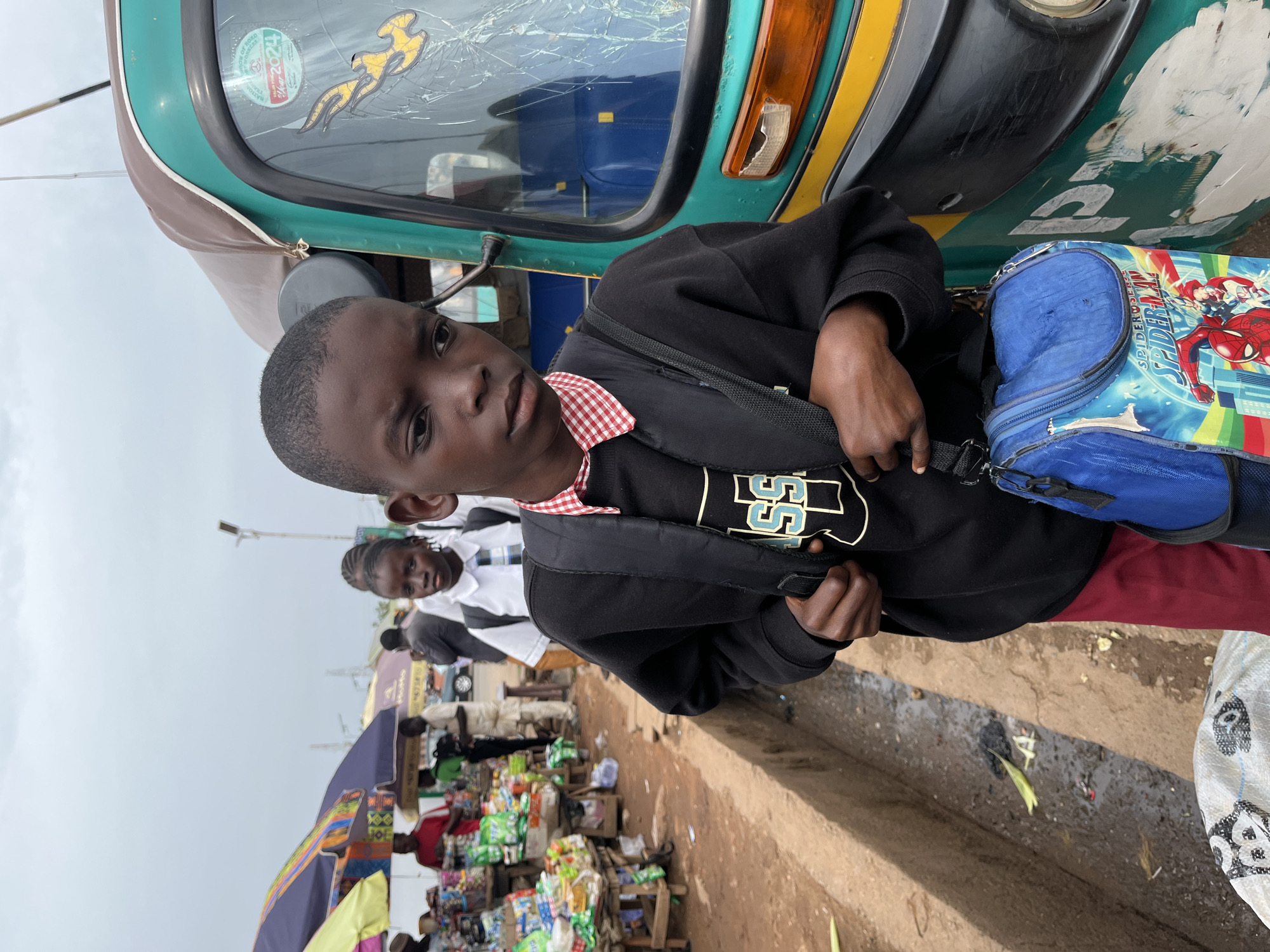 A young boy wearing a black shirt with a white and blue logo, a red and white checkered collar, and a blue Spider-Man backpack stands in front of a green and yellow auto rickshaw with a cracked windshield in Gura Top, Nigeria. Another person is standing behind the boy. A street market is visible in the background with vendors and their goods displayed on tables. The ground appears to be a mix of dirt and paved areas.