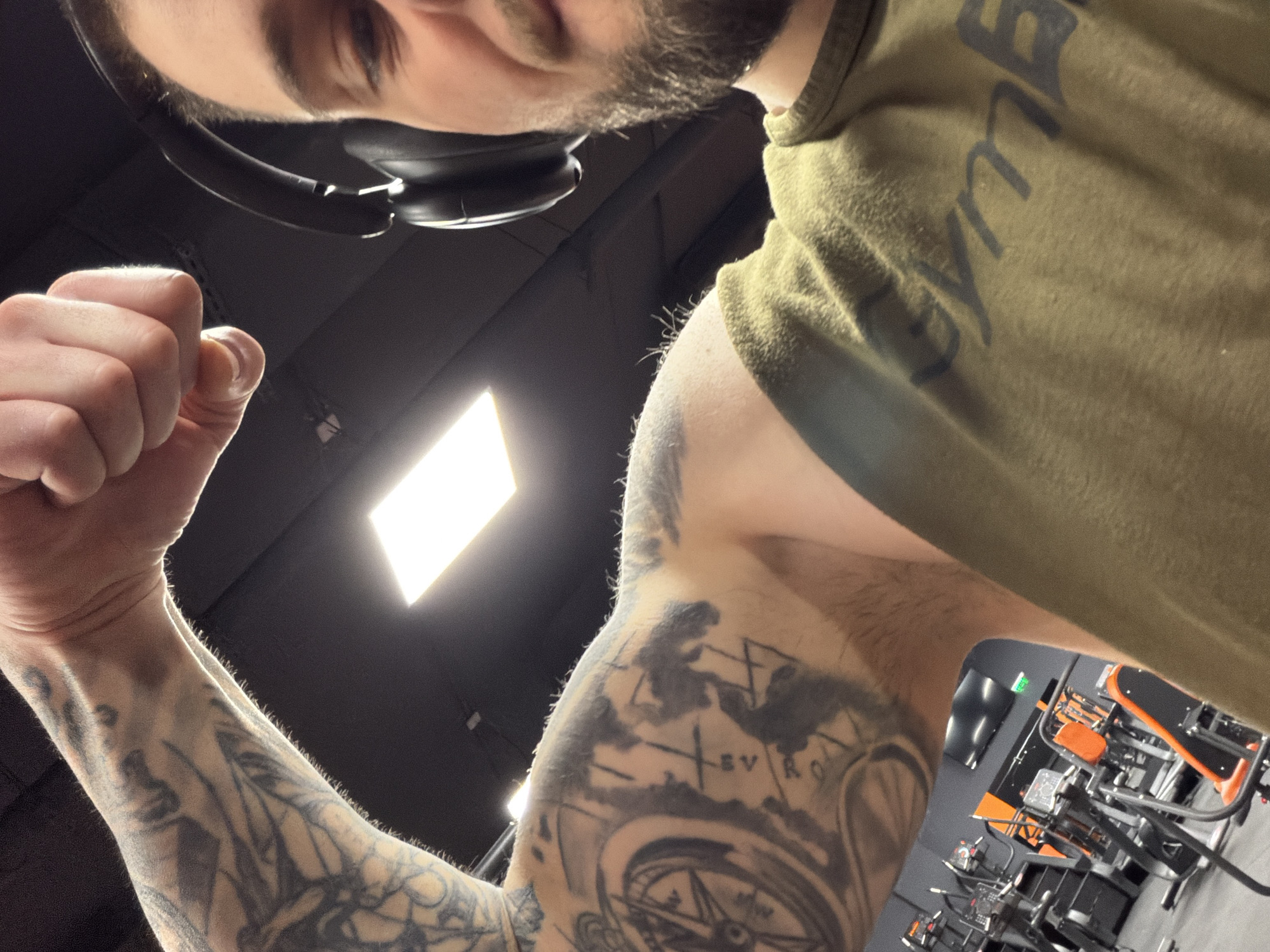 A man takes a selfie in a gym. He is wearing headphones and an olive-green t-shirt with writing on the front. The man is flexing his right arm, which has a tattoo featuring a compass and the word "EURO". In the background, gym equipment is visible. The gym is located in Plovdiv, Bulgaria, based on the tattoo featuring "EURO" as a reference. The gym appears to be in an area with dark ceilings and bright lighting.