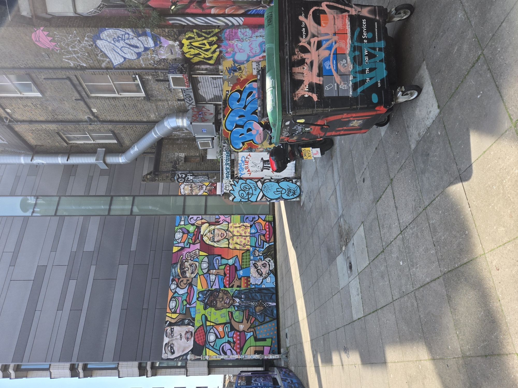 The image shows a street scene in London, United Kingdom. The left side of the image showcases a building with a modern architectural design, contrasting with an older brick building behind it. The buildings are decorated with extensive graffiti and street art, including a large colorful mural featuring various faces and figures. On the right, a commercial waste bin covered in graffiti stands on a paved sidewalk. A parked motorcycle is visible near the bin. The overall environment is urban, with an emphasis on the vibrant street art.