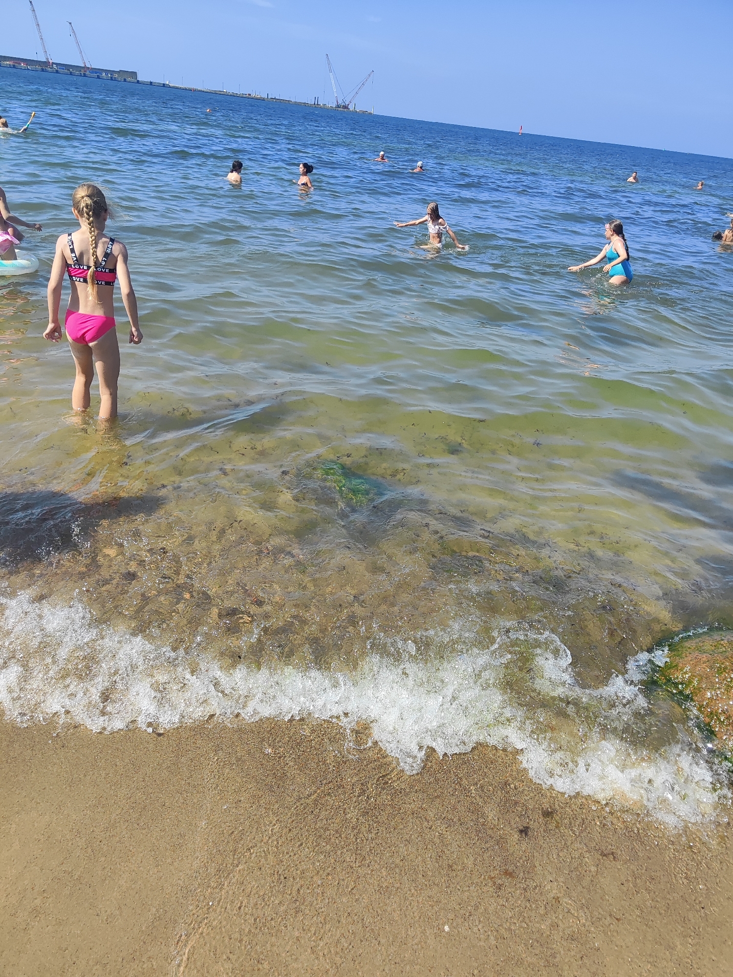 The image depicts a sunny day at a beach, likely in Pionersky, Russia, with several people swimming and playing in the shallow, clear water. In the foreground, a young girl with braided blonde hair stands facing away from the camera, wearing a pink bikini with "LOVE" printed on the top. The water is a mix of clear and greenish hues, with the sandy bottom visible beneath the surface. Small waves gently lap at the shore, creating white foam.
In the background, a diverse group of individuals, including children and adults, are scattered throughout the water. Some are wading, while others are swimming or engaging in playful activities. The sea stretches out to the horizon under a bright blue sky, indicating pleasant weather. In the far distance, industrial structures with cranes are visible along the coastline, suggesting proximity to a port or construction site. The overall atmosphere appears relaxed and recreational. The time of day is clearly daytime, given the strong sunlight and blue sky.