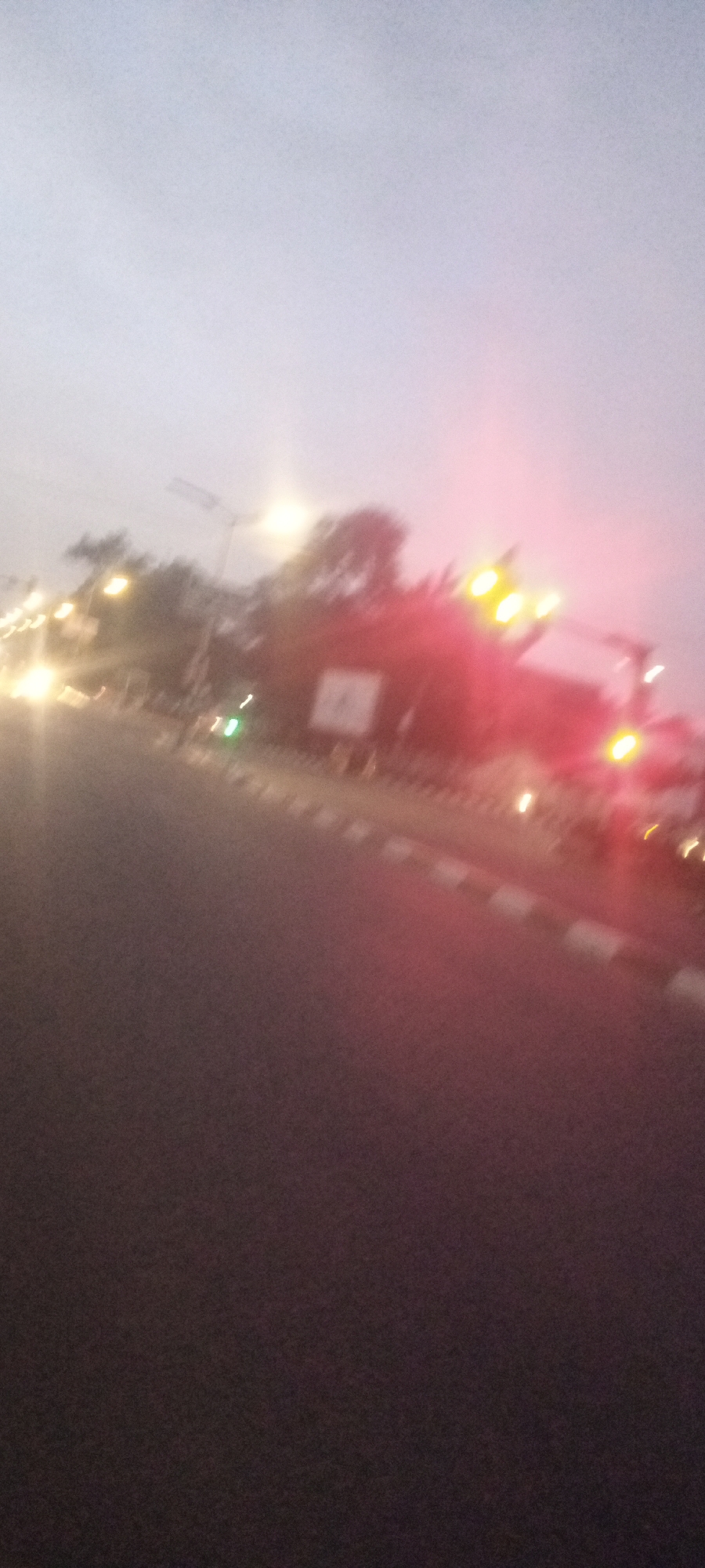 This blurry image shows a street scene, likely in Kabala, Nigeria. The foreground is dominated by the asphalt road. A curb with alternating white and dark sections runs along the edge of the road. In the background, there are streetlights, trees, and what appears to be a building or structure with lit red lights. There is also a sign with some text. The overall lighting suggests it is dusk or early evening. The lack of clarity makes it difficult to discern any other details with certainty.