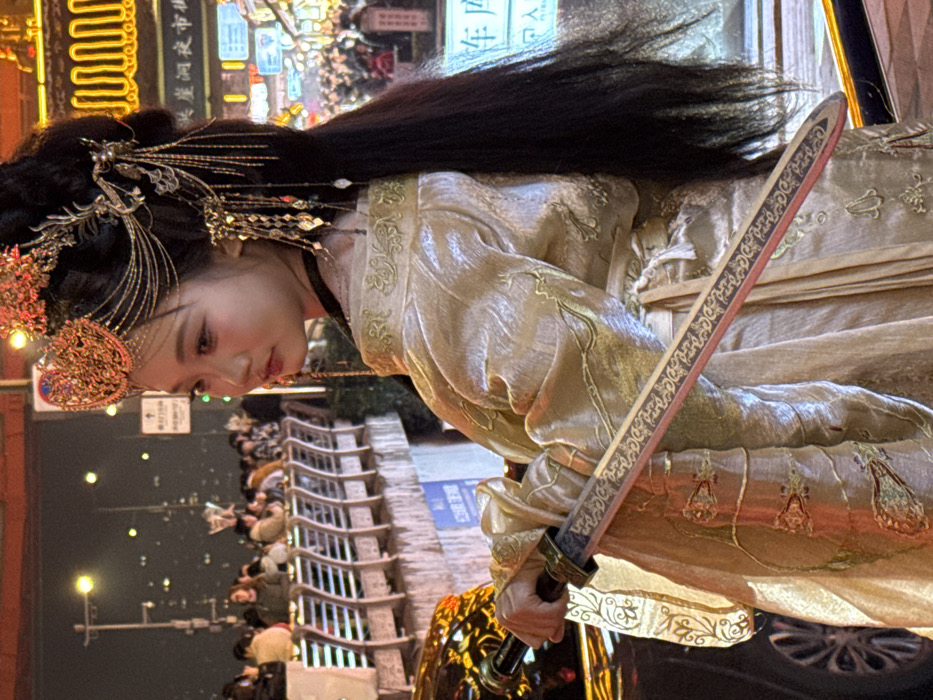 An outdoor, nighttime, medium-shot photograph taken in Yuzhong District, China, depicts a young East Asian female in traditional attire positioned left of center. She faces right, looking slightly away from the camera, with her body angled towards the left. Her long, dark hair is styled elaborately with a prominent gold-colored headpiece featuring a crown-like structure and multiple dangling chains and ornaments. She wears a shimmering gold-colored robe adorned with green and gold embroidery. Her left hand holds the hilt of a decorative sword, its dark scabbard with a red edge and gold patterns resting across her body.
The background is illuminated by artificial lights, indicating an urban setting. To the immediate left, a portion of a dark vehicle is visible. Further back on the left, a crowd of blurred individuals stands behind a light-colored metal railing, suggesting a walkway or bridge. To the right, a densely lit commercial street or night market is visible, with numerous lights and fragmented Chinese signage, including characters that appear to denote "night market" and "parking garage." Streetlights dot the dark sky in the distance. The overall scene conveys a lively, nocturnal city environment.