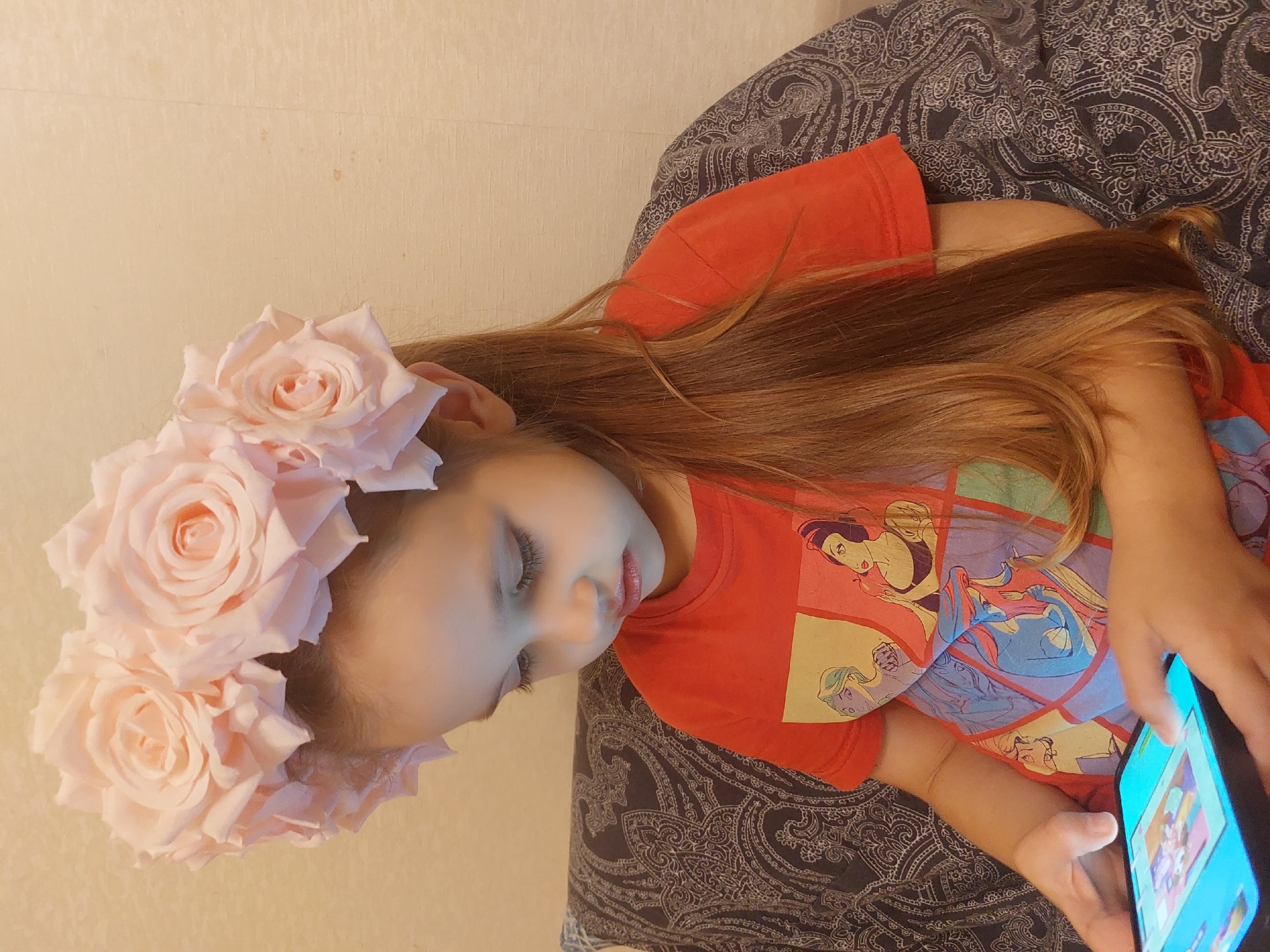 A young Caucasian female, with long brown hair, is sitting on a patterned sofa and looking down at a tablet. She is wearing a red t-shirt with an image of Disney princesses on it and a large floral headband made of light pink roses. The background is a textured wall. The image was taken indoors.