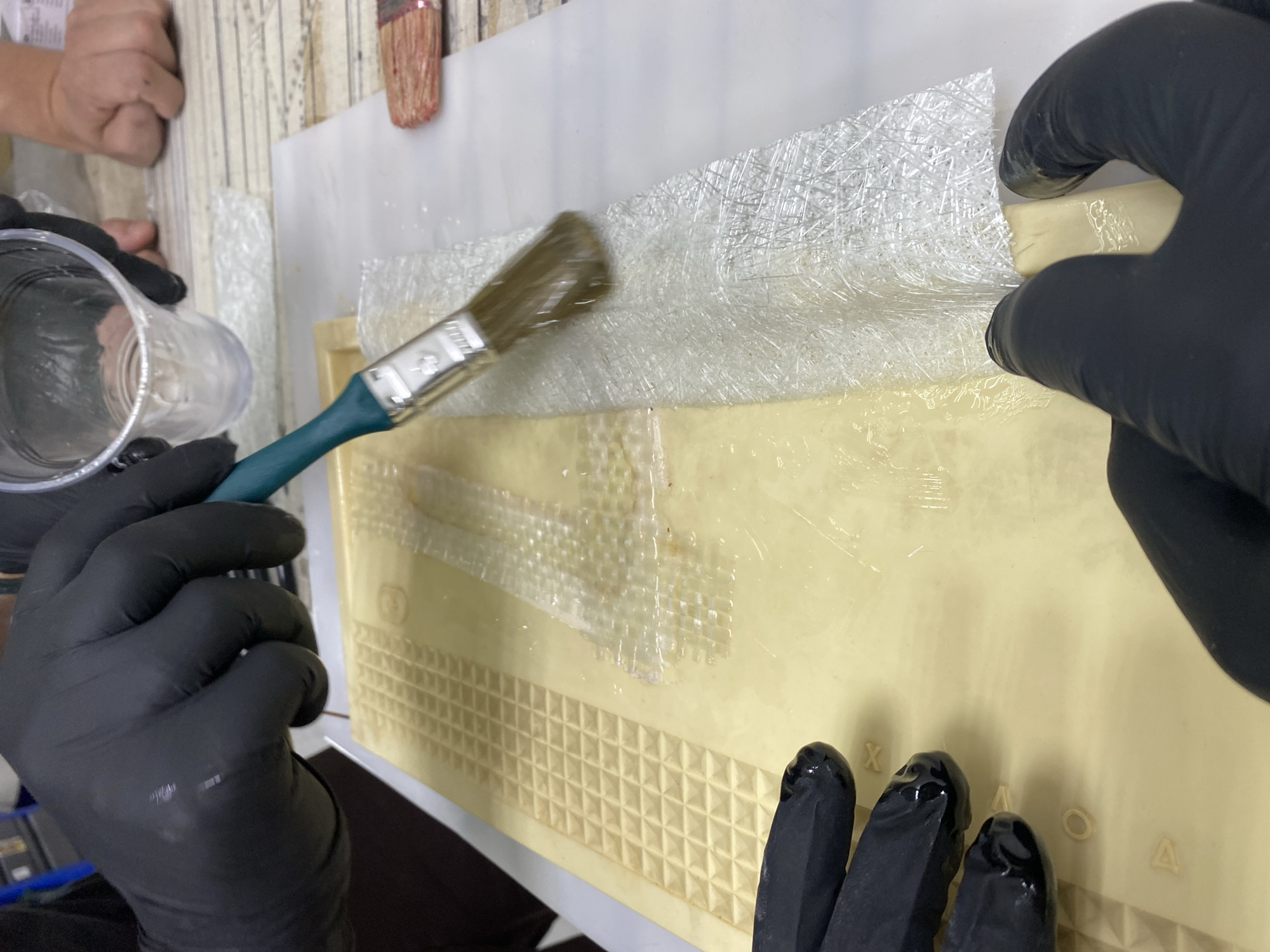 A first-person perspective shot shows a person wearing black gloves working on a fiberglass project. The subject is holding a clear plastic cup in their left hand and a paintbrush in their right, applying a liquid to a layer of fiberglass fabric spread over a light beige, textured surface. The fiberglass is translucent, with a woven pattern of fine threads. The liquid appears to be a resin, as it is clear and glossy, soaking into the fiberglass. The beige surface has an embossed grid pattern, with some areas taped off with clear tape. In the background, another person's hands are visible, also working on a similar project. A partially visible red-bristled paintbrush rests on a white sheet in the upper background. The lighting is bright, suggesting an indoor setting, possibly a workshop or studio. The scene conveys a sense of focused activity and crafting. There is no visible text in the image.