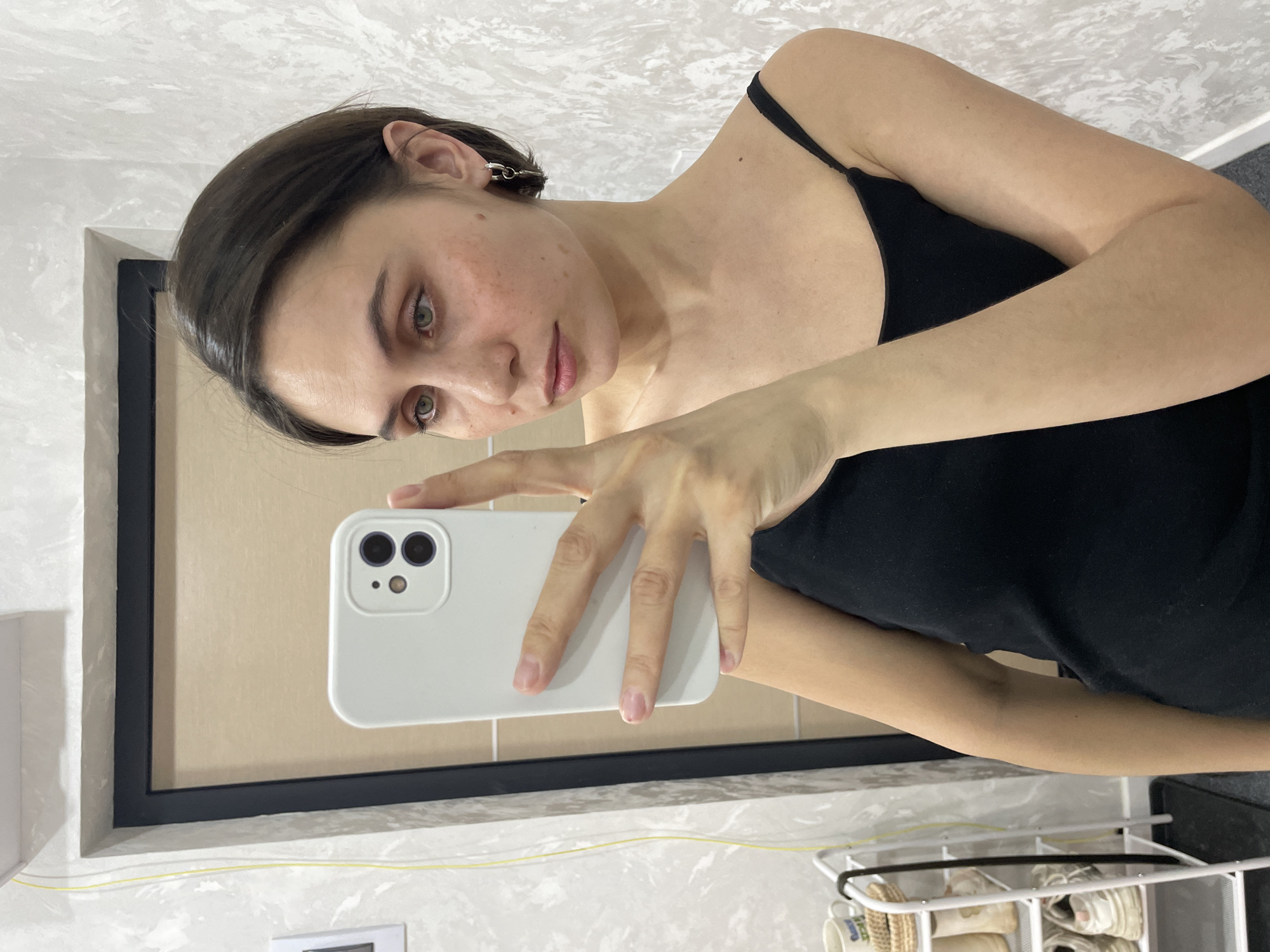 A young woman with short, dark brown hair, green eyes, and visible freckles is captured in a mirror selfie, holding a white smartphone. The image is oriented sideways, with the woman looking directly towards the camera reflected in the mirror. She is wearing a simple black spaghetti strap top and a single silver link-chain earring in her visible ear. Her right hand, with natural, short fingernails, holds the white phone which features a dual-camera system on its back.
The setting appears to be indoors, likely a bathroom or a dressing area. The background features light-colored tiled walls, some with a subtle marble-like pattern in gray and white. Behind the woman's reflection, the wall inside the mirror's frame is a plain light beige. The mirror itself has a slim black frame. Below the mirror, a thin yellow cable runs along the wall. Further down, part of a white metal shelving unit or shoe rack is visible, containing light-colored footwear and potentially a mug. The woman's expression is neutral and calm. The location of the photo is Izhevsk, Russia.
