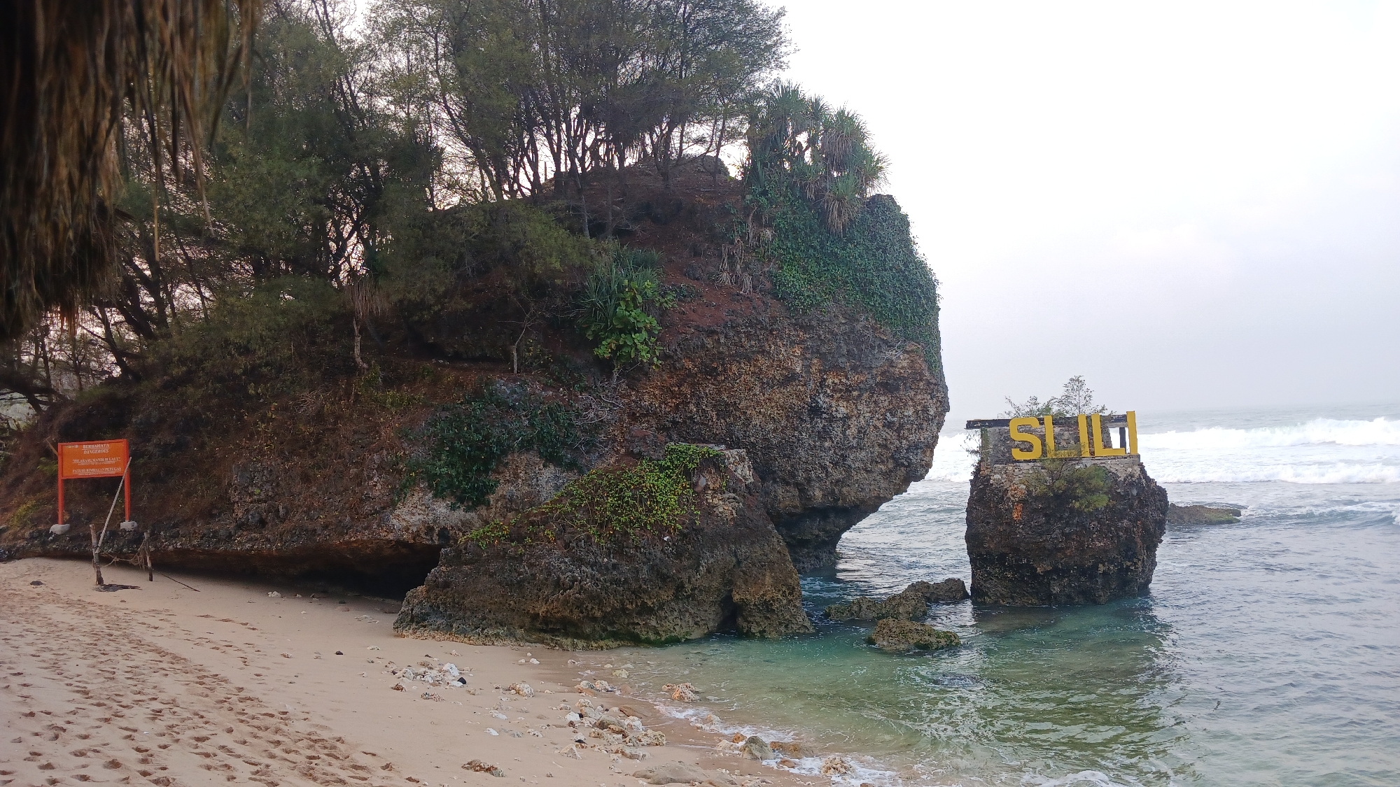 The image captures a serene coastal scene at Sili Beach, located in Tepus, Gunungkidul, Yogyakarta, Indonesia. The foreground features a sandy beach with footprints and scattered shells, leading to the clear, shallow waters of the ocean. On the left, a large, rocky outcrop covered with dense vegetation and trees rises from the sand. A prominent orange sign with Indonesian text, likely indicating safety warnings, is visible near the base of this outcrop. To the right, a smaller, isolated rock formation stands in the sea, topped with large, yellow letters spelling "SILI", serving as a landmark for the beach. Gentle waves lap against the shore and the rocks. The sky is overcast, casting a soft, diffused light over the landscape. The overall atmosphere is tranquil and natural.
