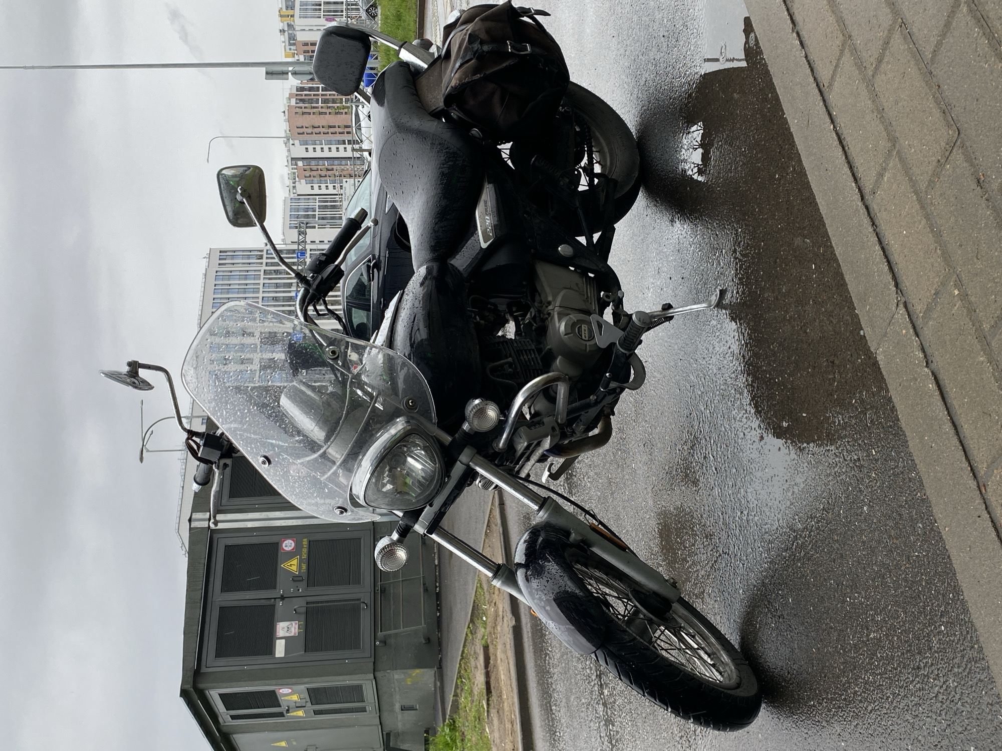 A black Bajaj Avenger 220 Cruise motorcycle is parked on wet pavement in what appears to be Saint Petersburg, Russia. The motorcycle has a windscreen, saddlebag, and the kickstand is deployed. Raindrops are visible on the windscreen and body. A grey electrical substation is on the left of the motorcycle. In the background are modern apartment buildings, consistent with architecture in Saint Petersburg. The sky is overcast, indicating recent or ongoing rain. The pavement is wet and reflective.