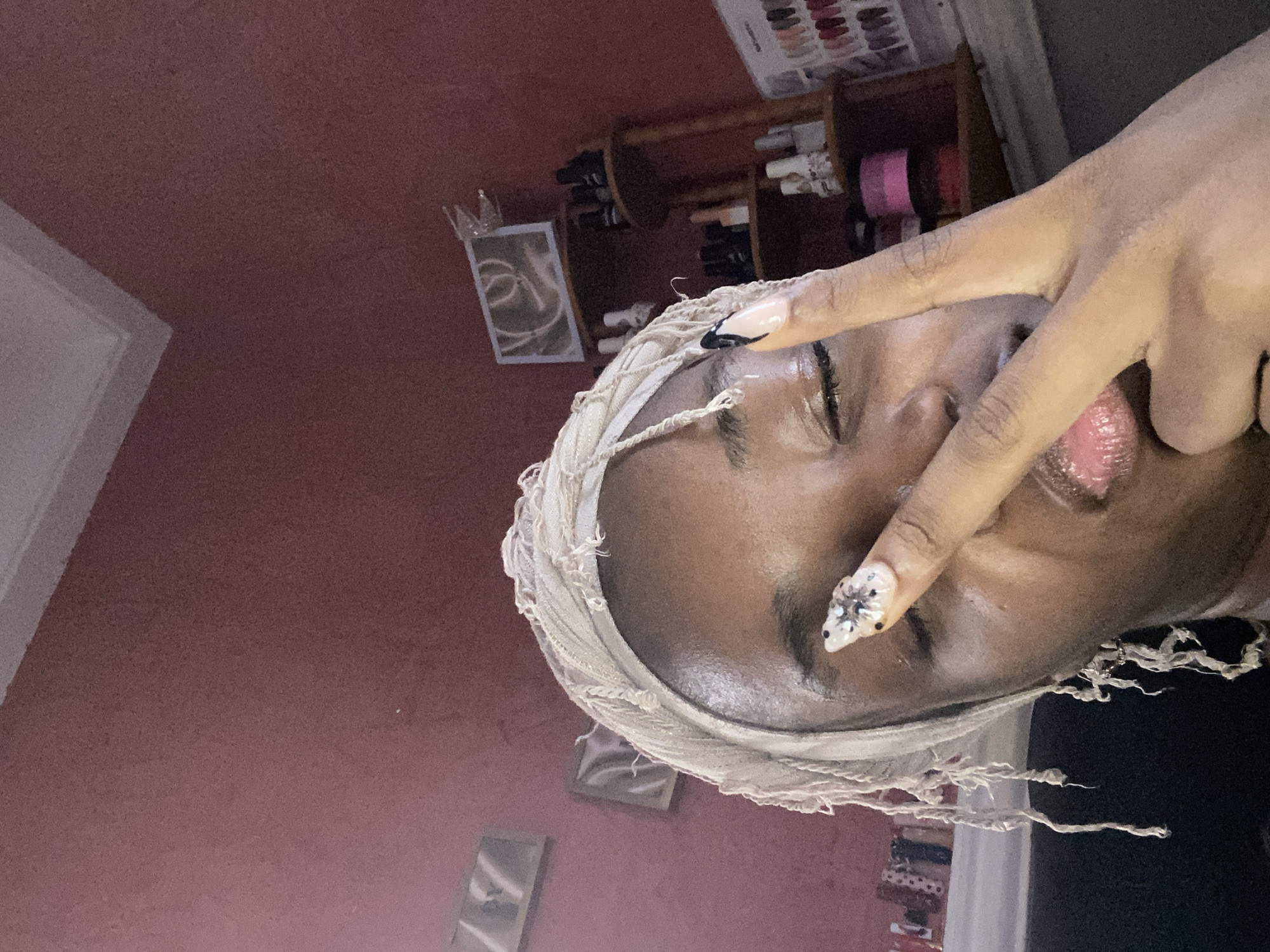 The image shows a close-up of a young Black person wearing a beige headscarf and making a peace sign with their right hand over their face. They have long, elaborate fingernails, with the visible nail adorned with a floral design and rhinestones. The person's eyes are partially closed, and they are wearing lip gloss. The background appears to be an indoor setting, with a pink-toned wall, a white ceiling molding, and shelves filled with various bottles, possibly nail polish or cosmetics, and framed pictures on the wall. One shelf contains several small bottles of what appear to be nail polish. Another shelf has a display of various colored nail tips. The overall lighting suggests an indoor environment, potentially a bedroom or a beauty salon. This image is not specific to Jos, Nigeria, as there are no identifying landmarks or contextual clues to suggest that specific location.