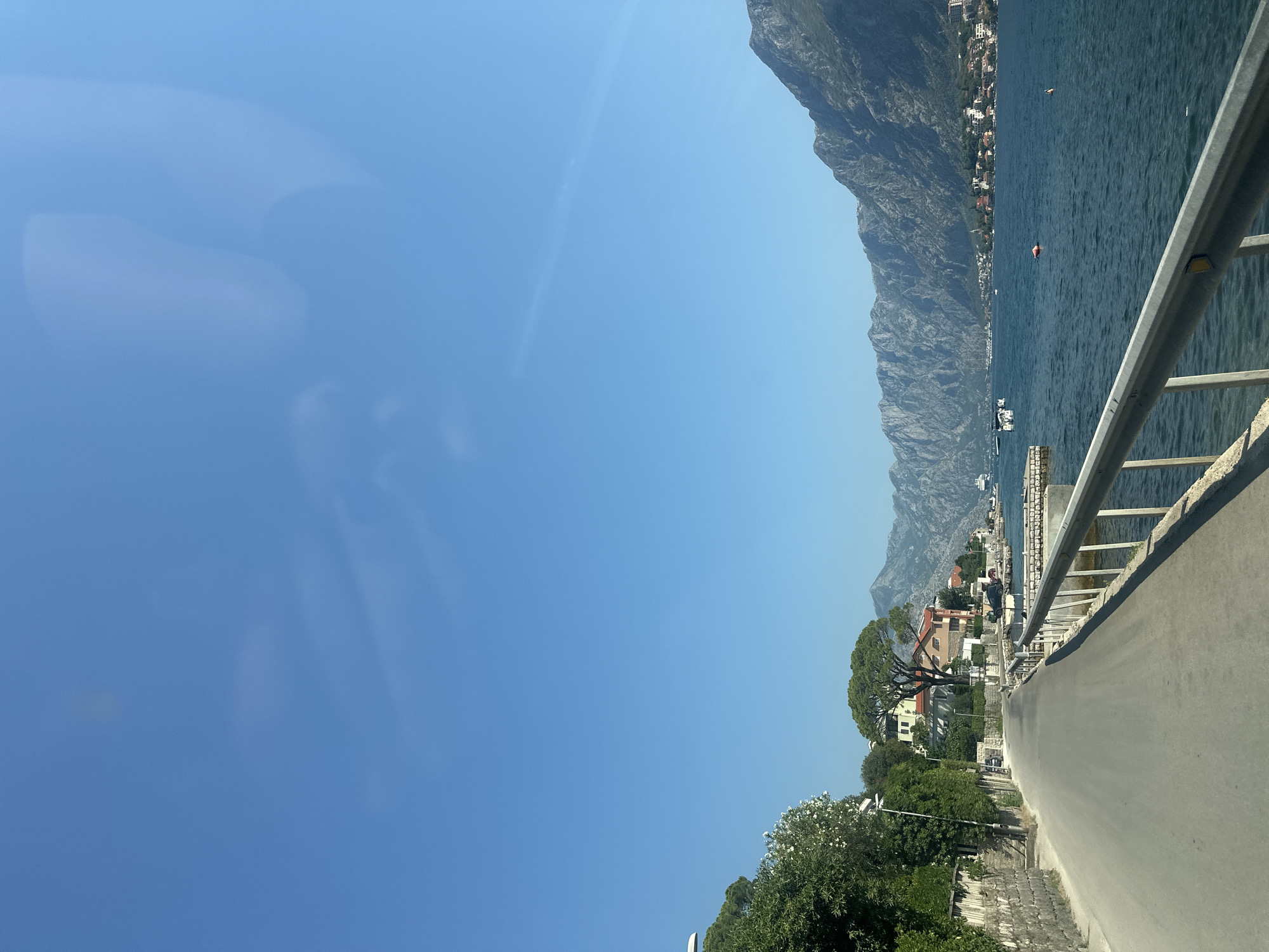 A sunny day at the Bay of Kotor in Muo, Montenegro, is depicted in this image. The foreground features a paved road curving to the left, bordered on the right by a metallic guardrail. Beyond the guardrail, the deep blue water of the bay is visible, with several small boats and a larger vessel in the distance. The left side of the road is lined with lush green trees and vegetation, with a stone wall at the very edge. In the mid-ground, a small cluster of buildings with red-tiled roofs are nestled amongst the trees, and a large, distinctive tree with a broad canopy stands prominently. The background is dominated by the imposing, rugged mountains that flank the bay, their slopes covered in vegetation and dotted with more buildings. The sky is a clear, bright blue with a few wispy clouds.