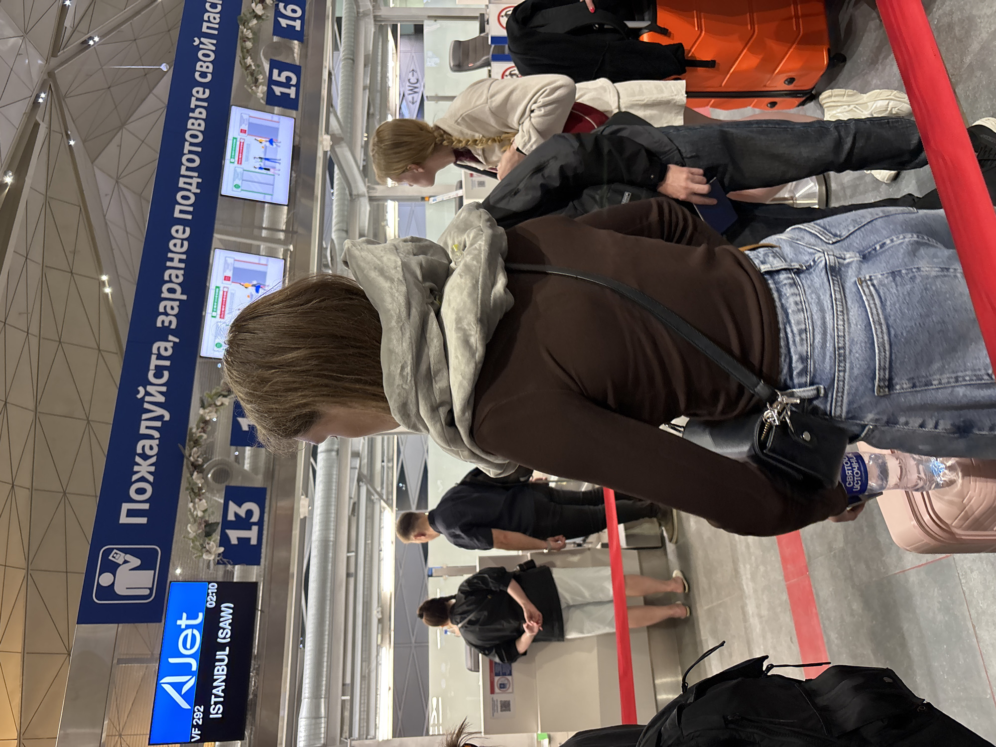 The image depicts an airport check-in area at Pulkovo Airport (LED) in Saint Petersburg, Russia. This identification is supported by the distinctive white grid ceiling with integrated lights, Cyrillic signage, and a visible "СВЯТОЙ ИСТОЧНИК" (Svyatoy Istochnik) branded water bottle. An overhead blue sign reads, "Пожалуйста, заранее подготовьте свой паспорт" (Please prepare your passport in advance). Below this sign, check-in counters numbered 13, 15, and 16 are present. A digital display indicates an "AJet" flight VF 292 to ISTANBUL (SAW), with a departure time of 02:10.
In the immediate foreground, a woman with brown hair, wearing a grey neck pillow over a brown long-sleeve top and light blue jeans, stands with her back to the camera. She holds a clear plastic "Svyatoy Istochnik" water bottle, which rests on a light pink hardshell suitcase beside her. To her right, partially obscured, a blonde woman with a single braid, dressed in a light beige jacket and white shorts, stands facing away from the viewer.
Further back, at one of the check-in counters, a woman with dark hair, wearing a black short-sleeve top and light-colored shorts, stands with her hands clasped behind her back. Beside her, a man with short dark hair, wearing a dark short-sleeve shirt and dark pants, faces the counter. The queuing area is delineated by a red barrier tape across the grey tiled floor. A "WC" sign is visible in the background, indicating restrooms.
