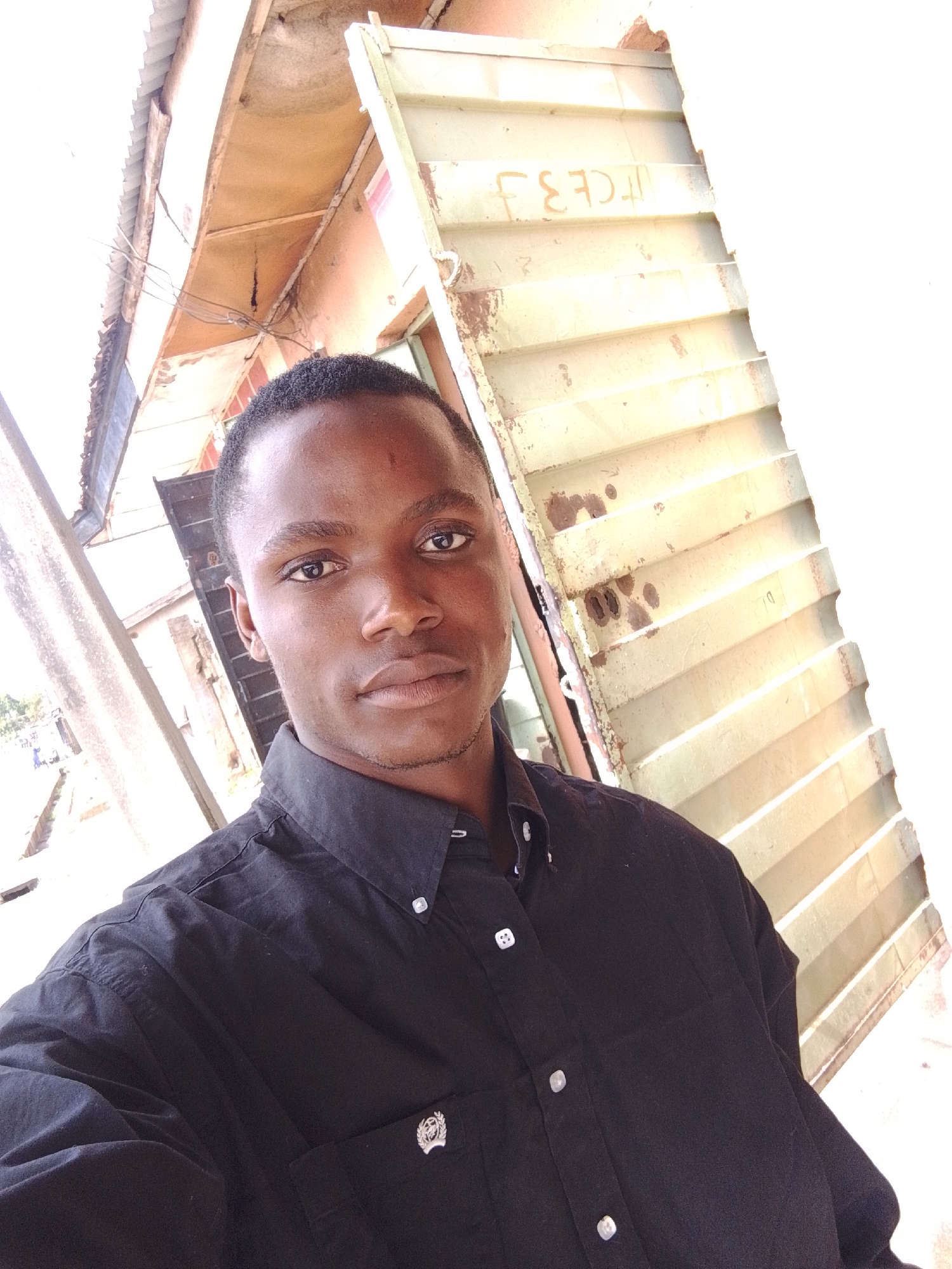 In Jos, Nigeria, a young Black man takes a selfie outdoors. He is wearing a black button-down shirt with white buttons. The man's head and shoulders are visible, and he is looking directly at the camera. The background consists of a corrugated metal structure, possibly a fence or wall, with some writing on it. The writing appears to be in orange and reads "ICE 3 J". The overall scene suggests an informal setting, possibly a residential or commercial area in Jos.