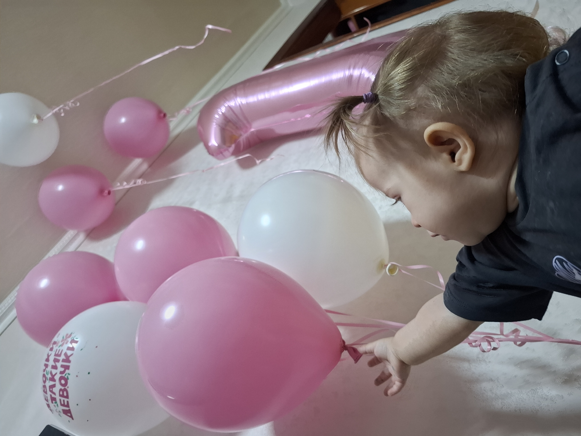 A young child with blonde hair pulled back in a small ponytail is reaching towards a pink balloon. The child is wearing a black shirt. Several other pink and white balloons are nearby, some with writing on them. The location appears to be indoors, possibly in a residence in Chaikovsky, Russia.