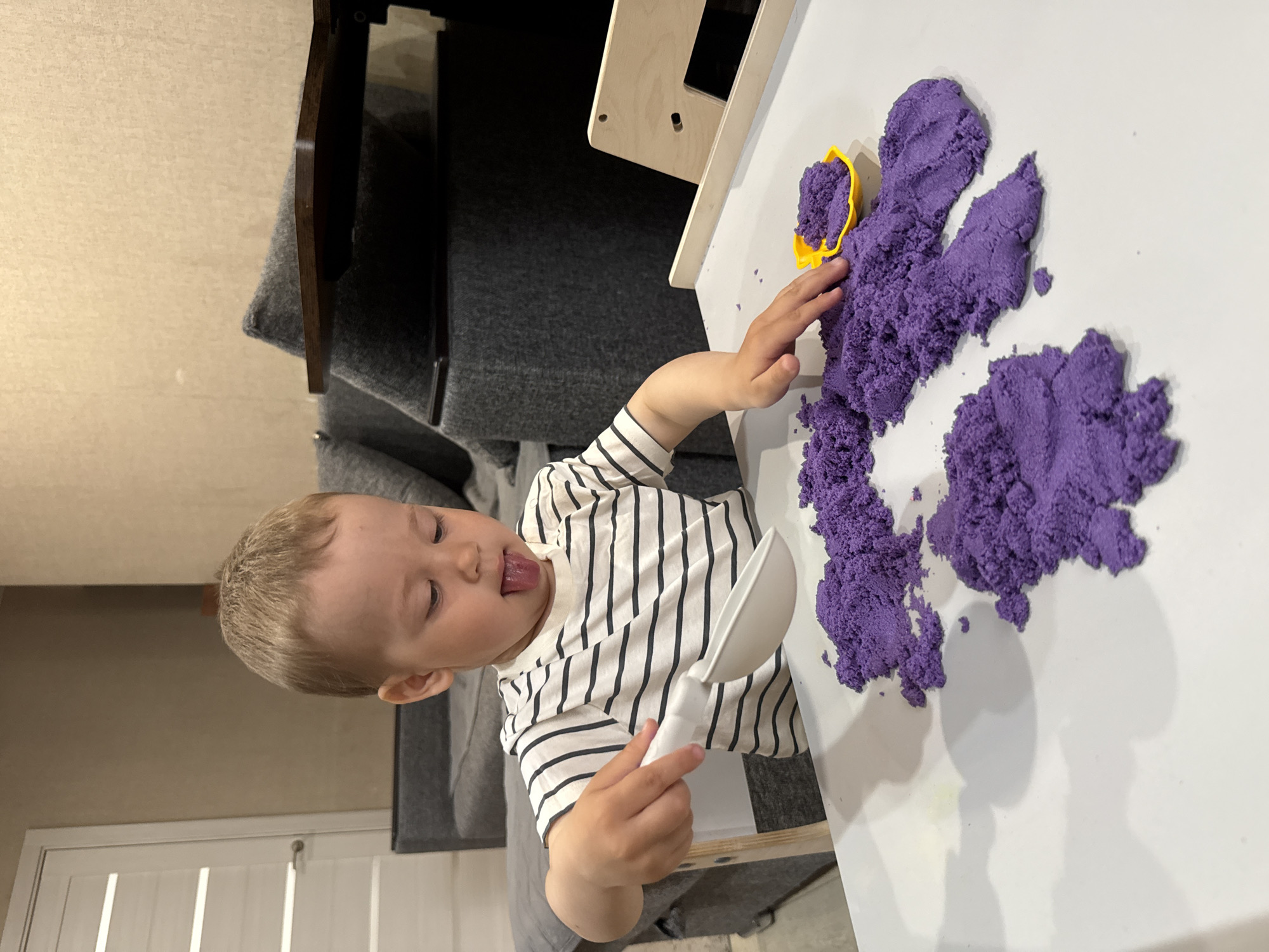 A young child with fair hair and a black and white striped shirt is playing with purple kinetic sand on a white table. The child is holding a white plastic scoop in one hand and touching the sand with the other. A yellow plastic mold filled with sand sits on the table next to the child's hand. Behind the table, there's a gray sofa, a wooden chair and possibly a children's ladder. No information indicates the location being Rusanovo, Russia.