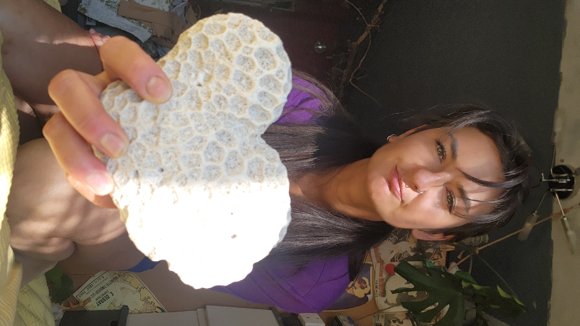 A person is holding up a light-colored, porous rock, possibly coral, close to the camera. The individual has dark hair, fair skin, and is wearing a purple top. They appear to have a nose ring and earrings. The background is cluttered, with items like a plant, books or papers with printed images, and what looks like a dark wall or curtain. The image is lit unevenly, creating harsh shadows on both the person and the rock. The OCR text suggests a connection to Paris, France, with references to "E. DEBRAY" and "PARIS." There is no indication of Moscow, Russia.