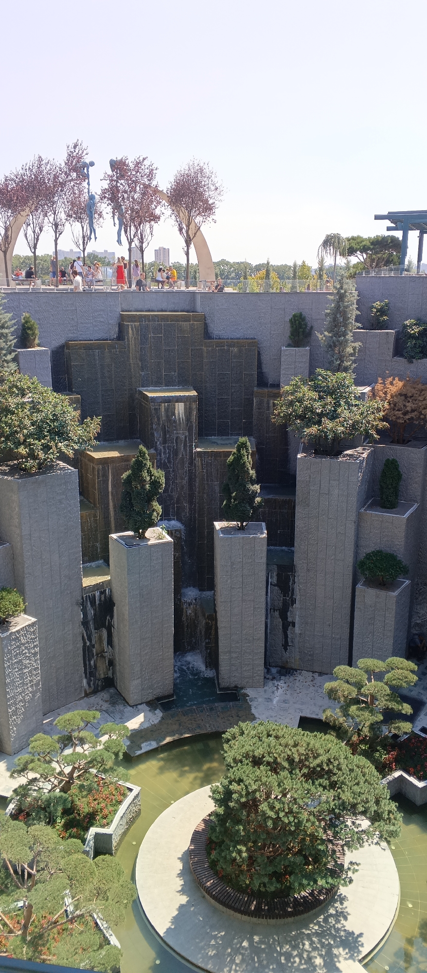 In an urban park in an unknown city, a tiered water feature made of stone cascades water down various levels. Tall, rectangular stone pillars are interspersed throughout the water feature, some topped with small trees. In the foreground, a circular concrete platform in a shallow pond features a large, mature evergreen tree with a wooden bench encircling its base. To the left, a planter bed is filled with smaller evergreen trees and red-leafed plants, surrounded by water. In the background, a crowd of people are visible on an elevated walkway, with trees and distant buildings under a clear sky.