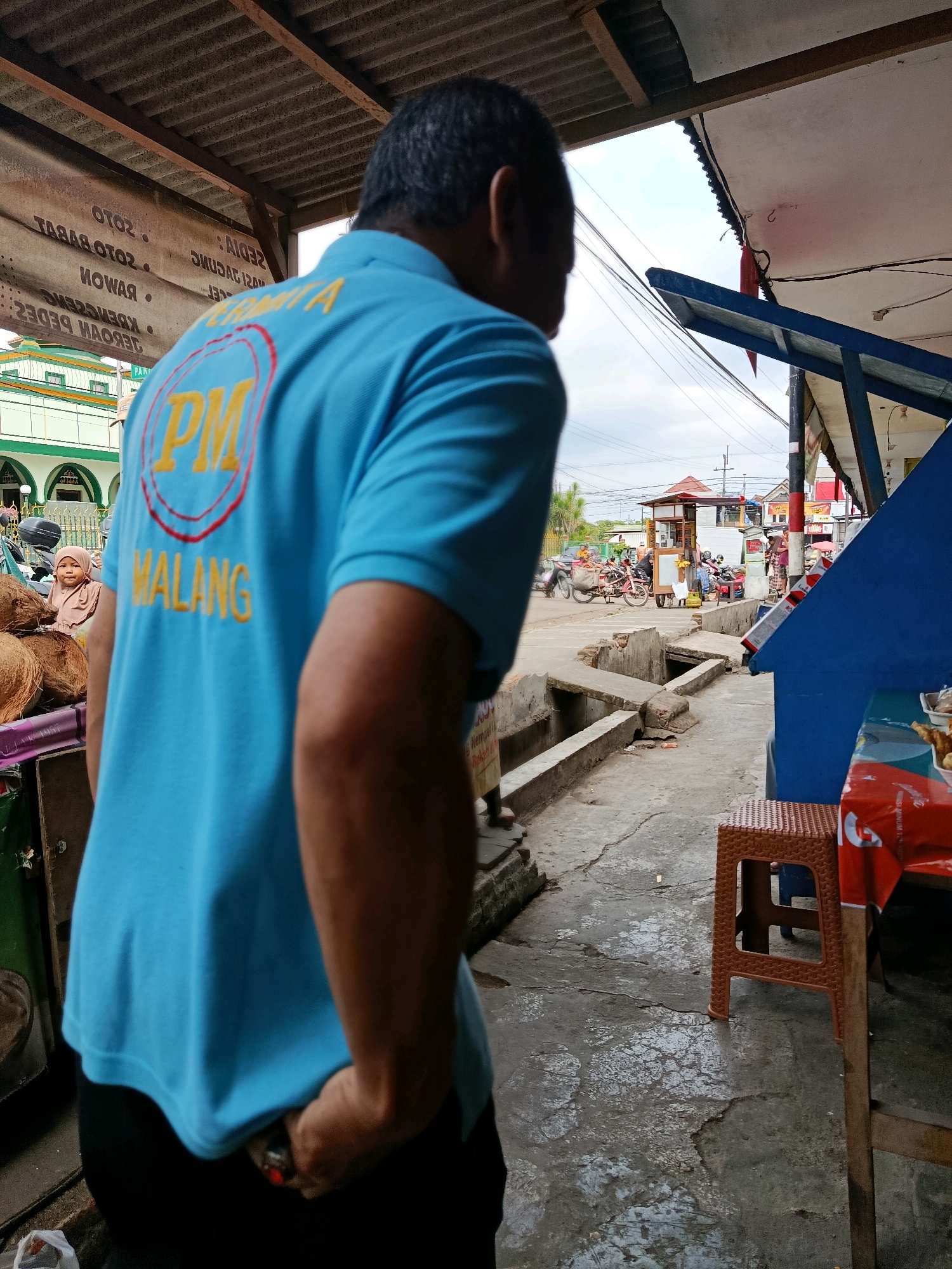 The image depicts an outdoor scene in what appears to be a market or street-side area in Bululawang, Indonesia. In the foreground, a person wearing a bright blue polo shirt with yellow lettering is seen from behind. The shirt has a circular logo with "PM" in yellow letters. The person's right hand is in their pocket. To the left, a stall or market display is partially visible with coconuts. In the background, a mosque with green domes and white minarets is situated. Further back, there are other buildings, street vendors, and various vehicles including motorcycles and a tricycle. The ground is paved with concrete, showing some wear and tear, and a drainage channel runs along the side. The sky is cloudy.