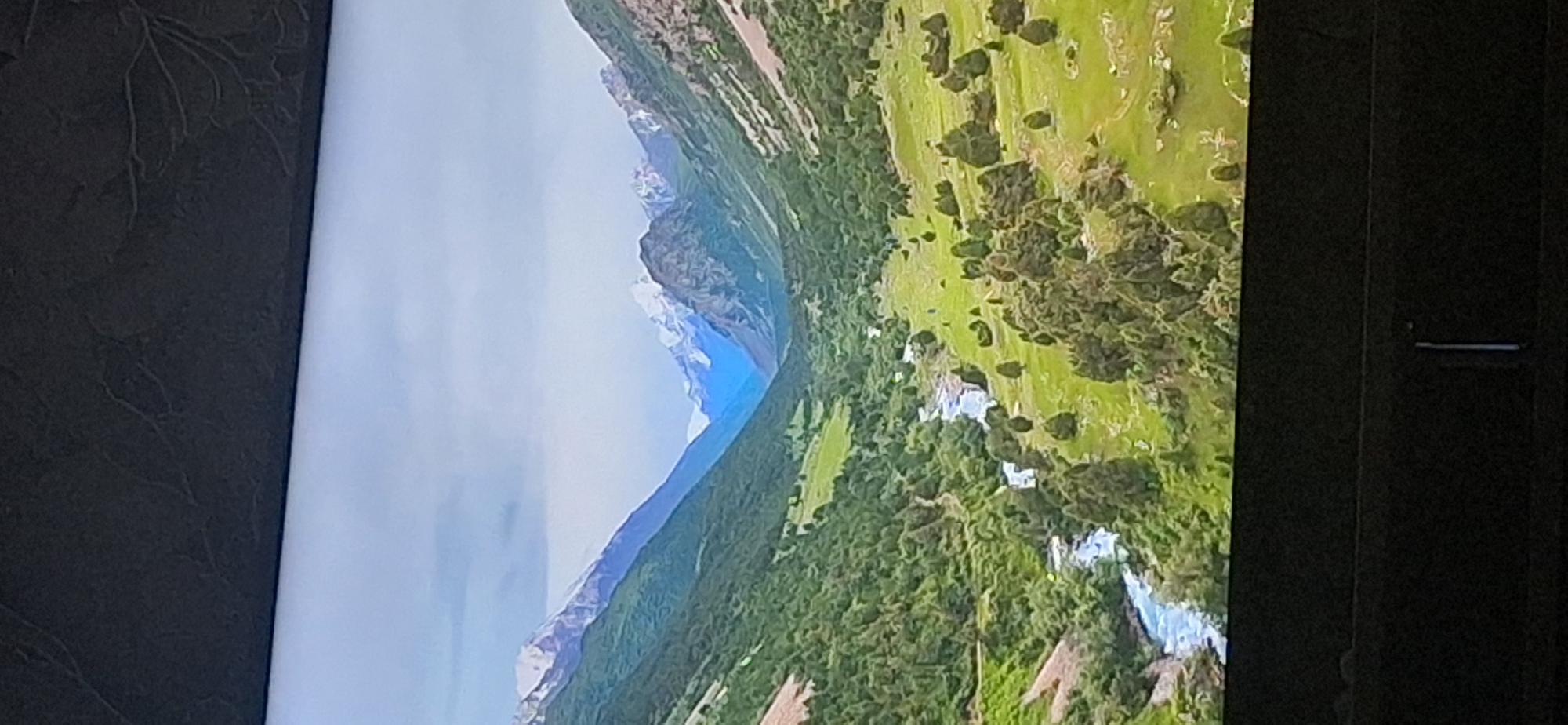 This image depicts a scenic mountain valley with a river flowing through it. The valley is filled with lush green trees and meadows, while snow-capped mountains rise in the distance under a cloudy sky. The perspective is from a high vantage point, looking down into the valley. The scene is entirely natural and does not contain any elements that can be identified as being located in the city of Bila Tserkva, Ukraine.