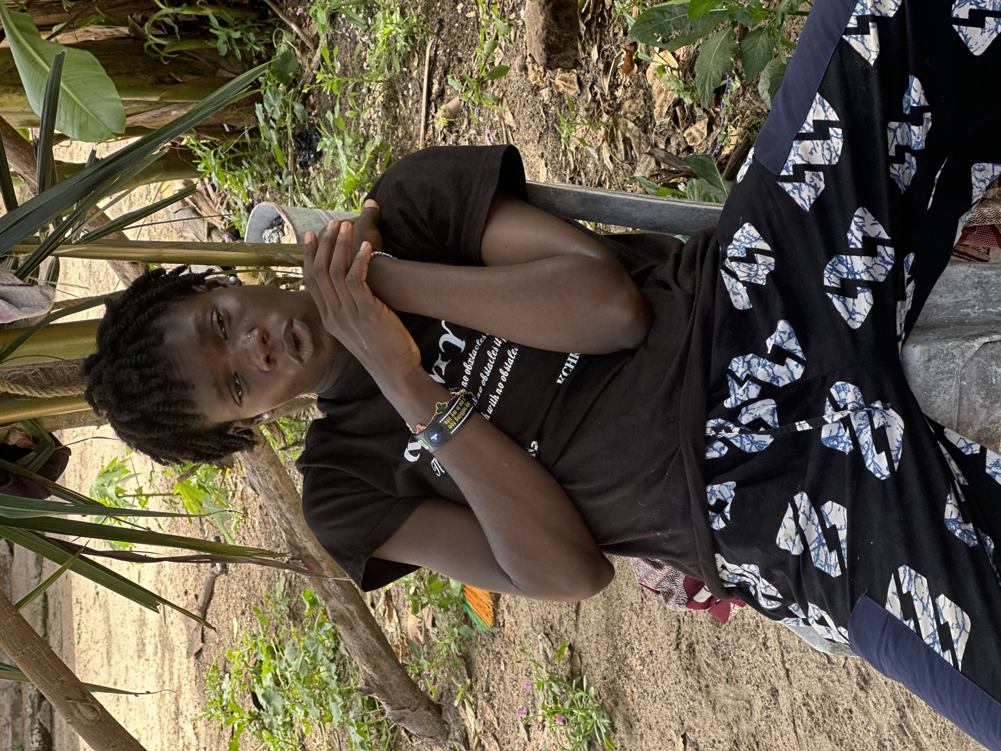 A Black individual with dark, coiled hair is seated outdoors in a sandy area, possibly within the city of Tawpo, Nigeria. They are wearing a black t-shirt with white text and black pants adorned with a blue and white pattern. Their arms are crossed, and they are looking slightly to the left with their mouth open. The background features vegetation, including banana trees and other plants, along with branches and some debris. A metal pole is visible to the right of the individual, and a bucket can be seen behind them. The lighting suggests it is daytime.
