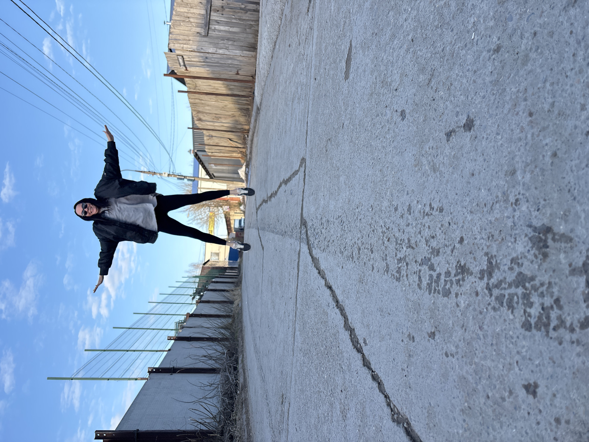 A woman is captured in a full-body shot, positioned at an oblique angle, seemingly suspended against a backdrop that includes a clear blue sky. She is dressed in a black jacket, a grey shirt, and black leggings, and wears sunglasses. Her arms are outstretched to the sides, and her legs are spread apart in a wide stance.
The environment includes a concrete surface that occupies a significant portion of the foreground, suggesting a street or sidewalk. Along the edges of the concrete, there is a fence-like structure and a wooden fence, indicating an urban or semi-urban setting. Overhead, power lines stretch across the sky. Based on image analysis and available geotags, it is plausible the location is in the city of Chaikovsky, Russia.