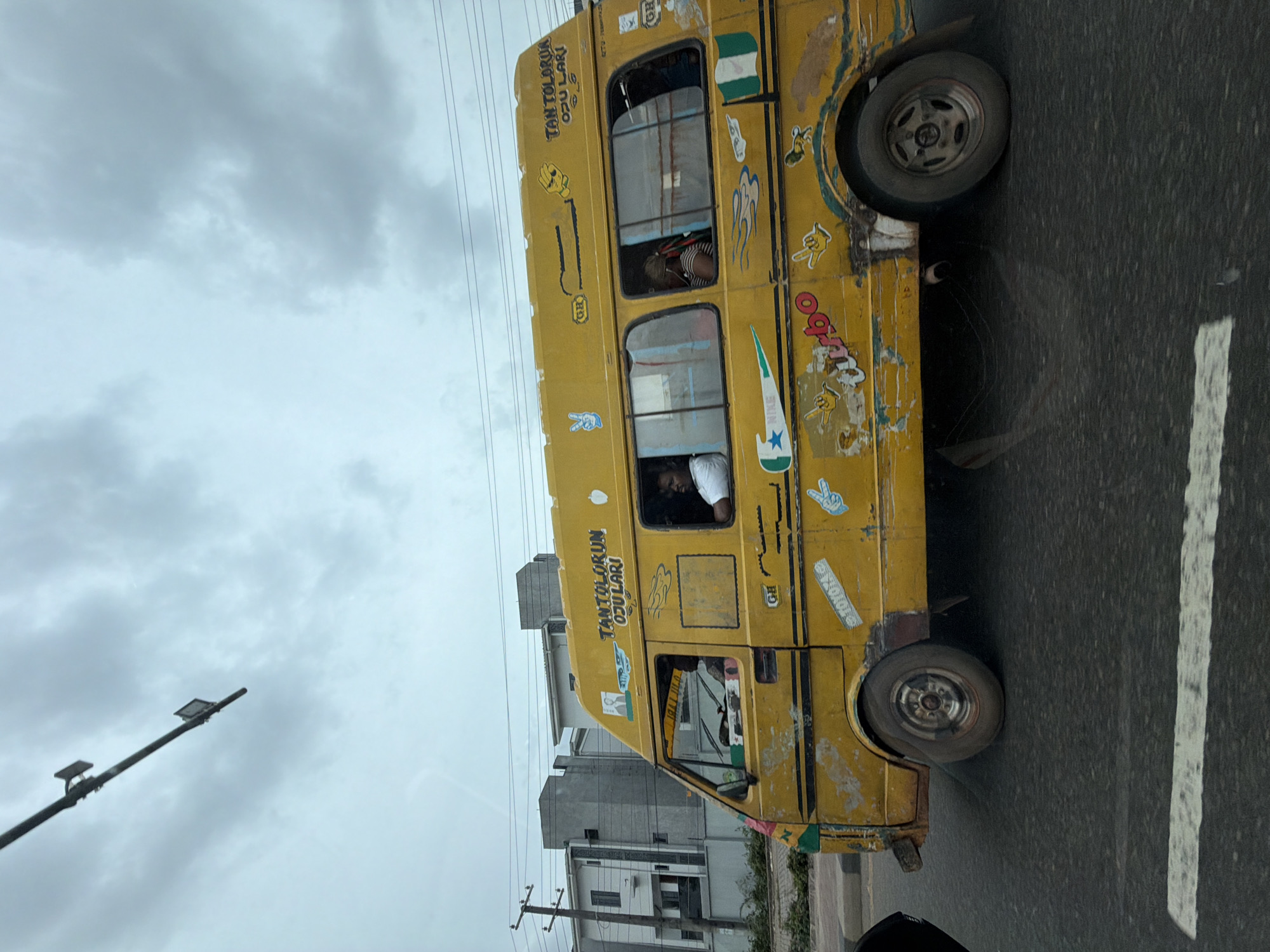 The image shows a yellow minibus, commonly known as a "danfo," on a road in Ikeja, Nigeria. The minibus has the inscription "TANTOLORUN OJULAR" painted on its side. It is adorned with various stickers and decals, including the Nigerian flag, and appears well-worn. Passengers are visible inside the vehicle. The minibus is on a road with visible lane markings. In the background, there are buildings, utility poles with power lines, and a cloudy sky. A streetlight is also visible in the frame. The scene seems to depict typical urban transportation in Ikeja.