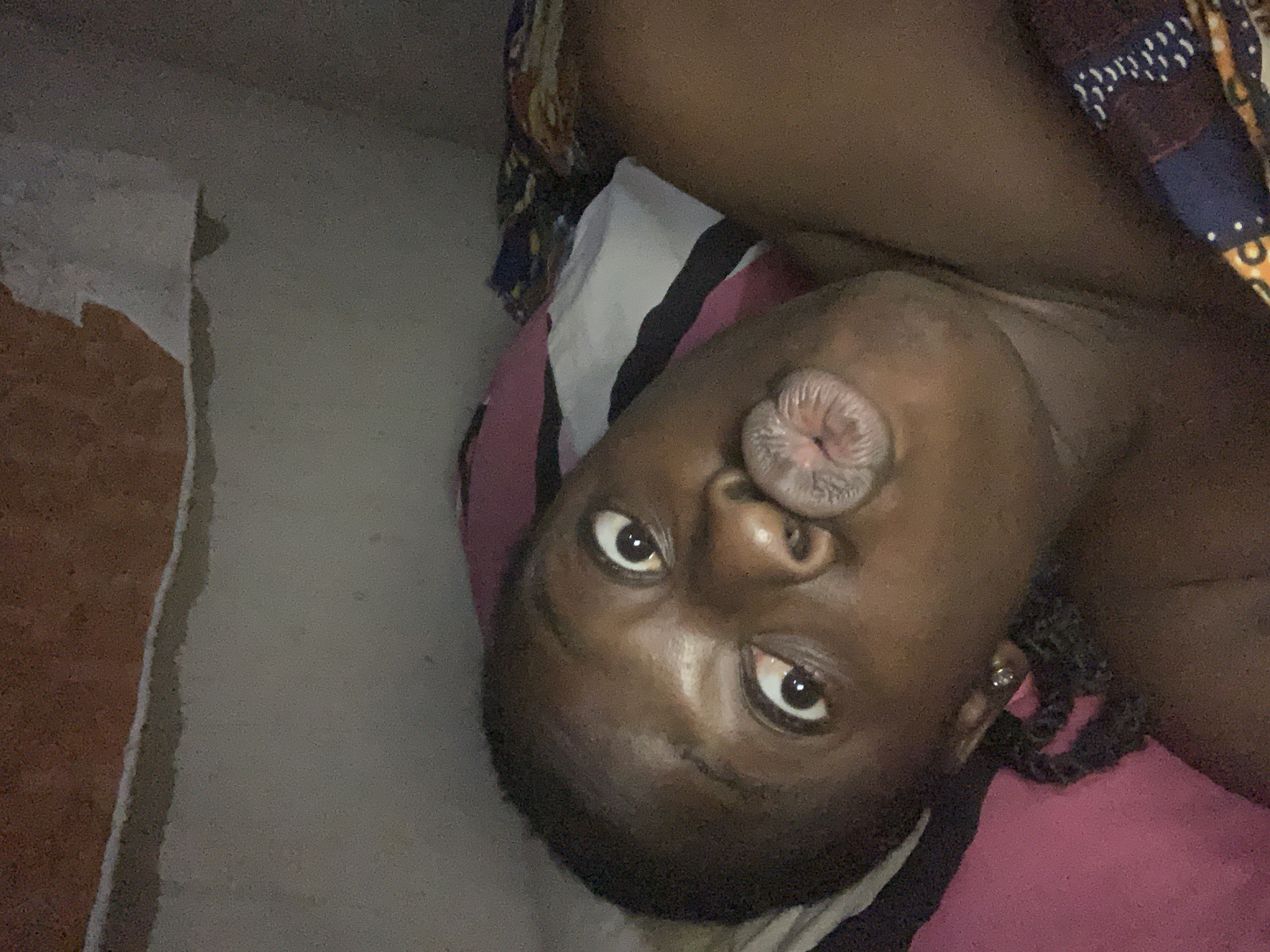 A dark-skinned person is lying on their back, appearing to be on a bed or resting on pillows with pink and white fabric. They are looking directly at the camera with their lips pursed in a kissing expression. Their head is tilted, and part of their shoulder is visible. The background includes a wall with damaged or peeling paint. The location is not verifiable as being in the city of Kukum, Nigeria, as that information is not discernible from the image.