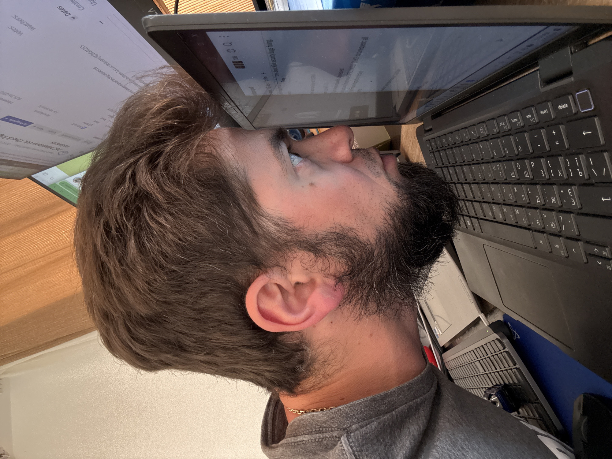 A person with light skin, a beard, and brown hair is looking at a laptop screen. They are wearing a grey shirt and a gold chain necklace. The laptop has a black keyboard. Another screen is visible on the left. The OCR suggests that the screens display "JBC Milestone GA: Check log" and some testing instances information. The image also contains words in what appears to be a keyboard layout containing Cyrillic alphabet.
There is no information in the image to suggest that it was taken in Šušanj, Montenegro. The OCR, the subjects, objects, and actions present do not provide conclusive evidence of location.