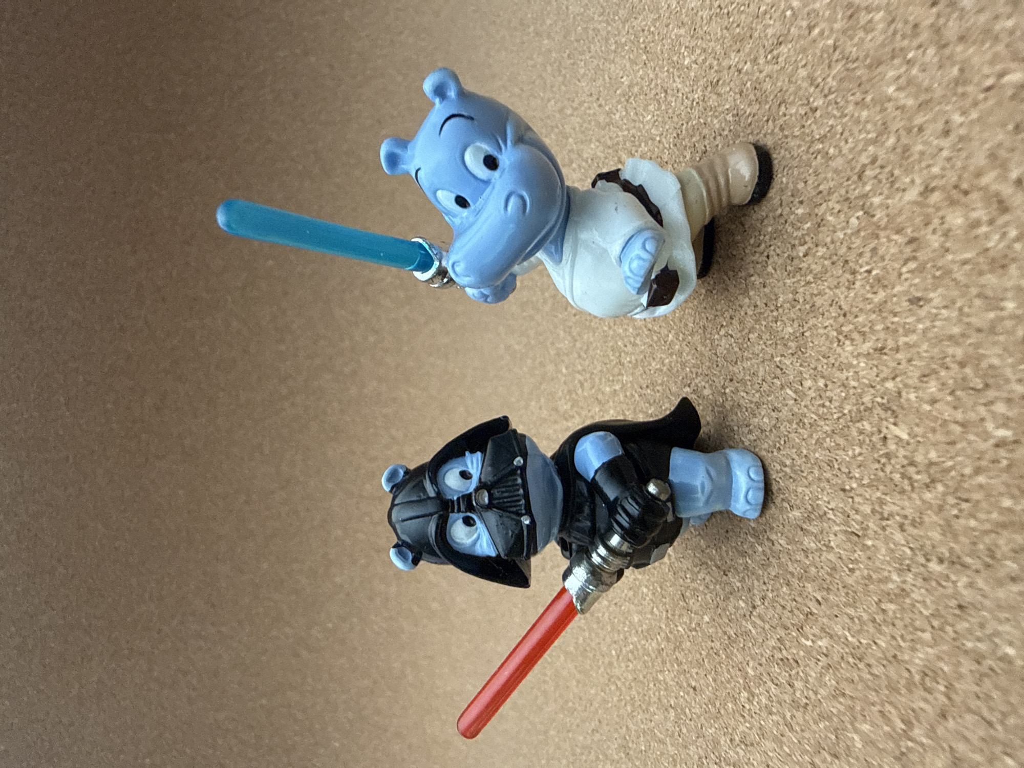 Two cartoon hippopotamus figures are lying on a light brown corkboard surface. The upper figure is light blue and dressed in white Jedi robes, wielding a blue lightsaber. The lower figure is dressed as Darth Vader in a black suit and mask and wielding a red lightsaber. The background consists solely of the corkboard surface.