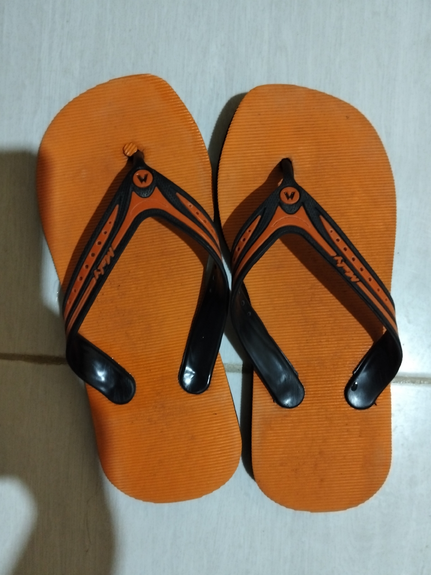 A pair of orange flip-flops with black straps is laid out on a tiled floor. The flip-flops are oriented with the soles facing upwards, and they are positioned side-by-side. Each strap features an orange accent with a black "W" logo and a pattern of small orange dots. The right flip-flop also has "Maly" written in orange script on the strap. The tiled floor appears to be a light beige or white color, and a shadow from the flip-flops is visible on the wall behind them. This scene is located in Payagaboh, Indonesia.