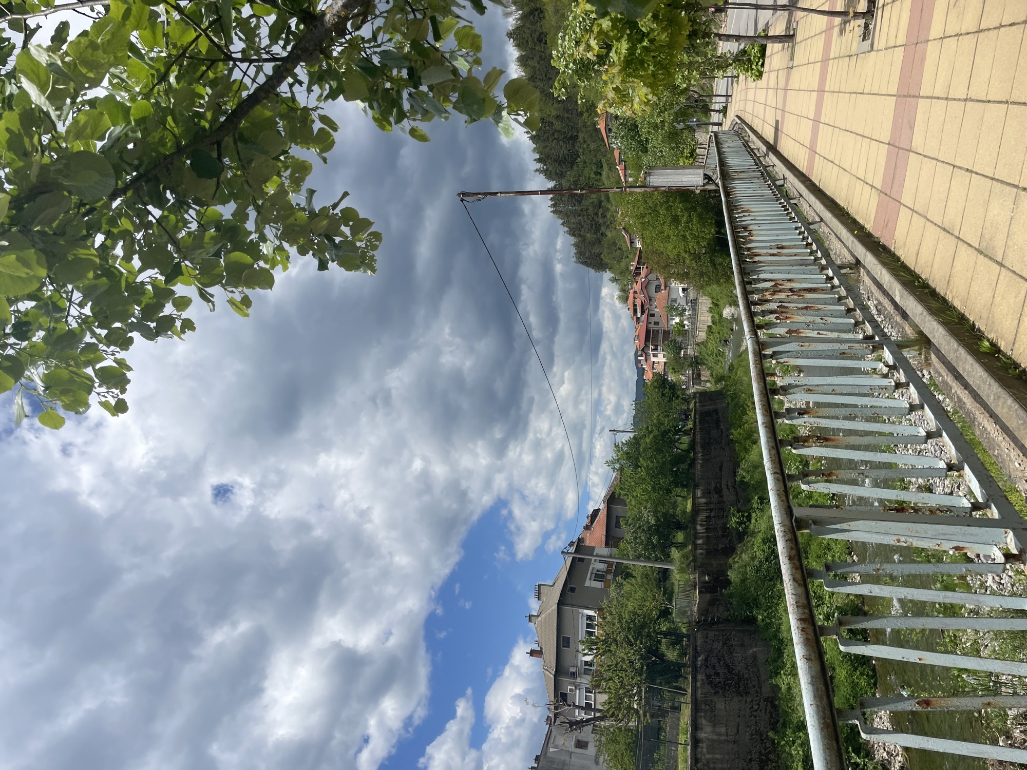 The image shows a view in Zlatograd, Bulgaria, featuring a sidewalk made of yellow and pink patterned tiles, a metal railing with rusty details, and a view overlooking a river valley. The foreground includes tree branches with green leaves. In the midground, there is a view of houses with red-tiled roofs and green trees. The sky is partly cloudy with a mix of dark and light clouds. An electrical pole and wire are also visible.