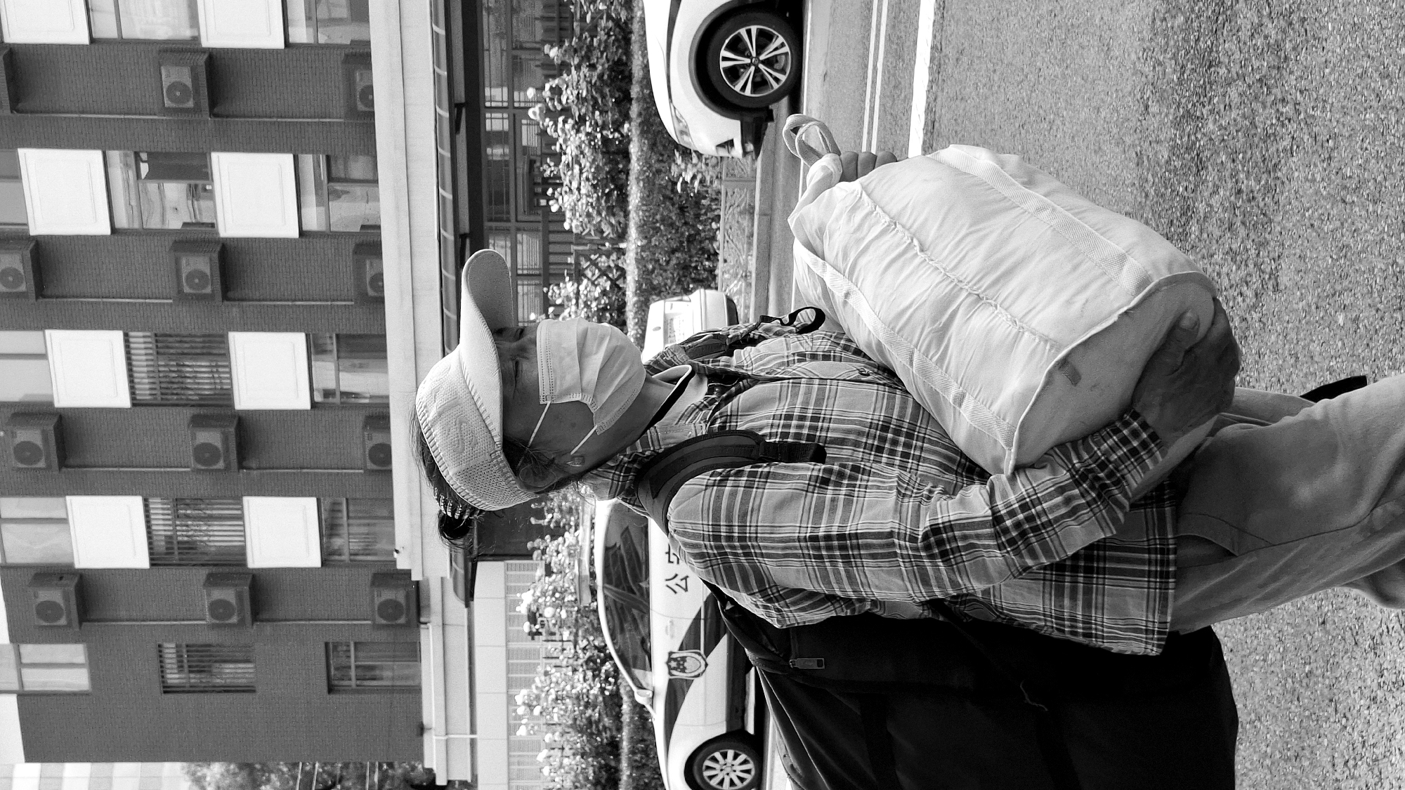 A grayscale medium shot shows a man carrying a large pillow on his shoulder, wearing a plaid shirt, a backpack, a hat, and a face mask. The man is standing on an asphalt surface, likely a street or sidewalk. Behind him, a building with numerous air conditioning units mounted on its exterior wall is visible. Cars are parked alongside a curb in the background. The environment appears to be urban, possibly in the Dongcheng District of China, given the architectural style and the possible Chinese characters on a vehicle.