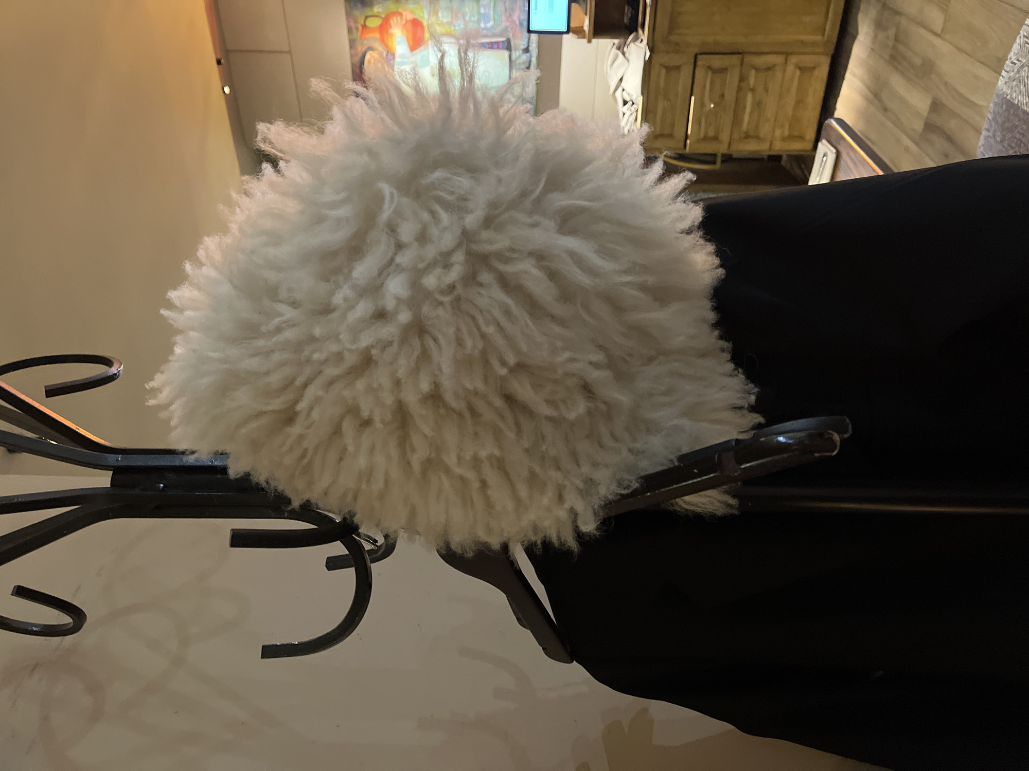 A fluffy white hat made of shaggy material sits on a black coat draped over a dark metal coat rack. The coat rack has several curved hooks. In the background, a wooden dresser with three drawers is visible. A laptop sits on top of the dresser, and a colorful painting hangs on the wall behind it. The floor is made of wood planks. The scene appears to be an indoor residential setting, possibly in Khimki, Russia.