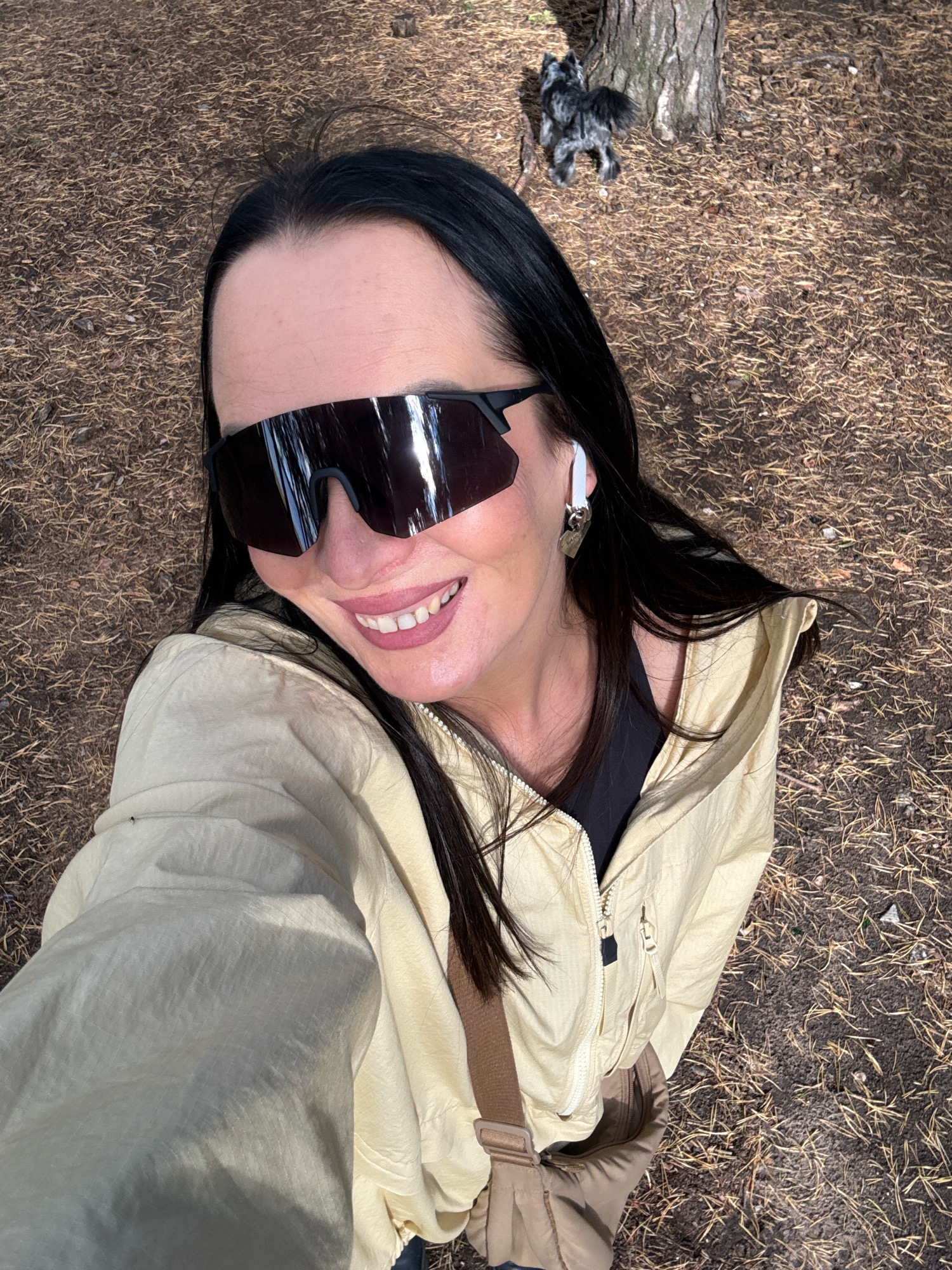 A selfie-style, medium shot captures a woman with long, dark hair and black, oversized sunglasses smiling at the camera. She is wearing a light yellow zipped jacket over a dark top and a tan crossbody bag. White wireless earbuds are visible in her right ear. The background consists of dry pine needles and soil, with a tree trunk and a small, dark-and-white dog in the upper right quadrant. The scene appears to be outdoors, potentially in a park or wooded area. The woman is positioned in the foreground, with the dog and tree further back. There is no identifiable landmark or context suggesting the specific location within Chaikovsky, Russia.