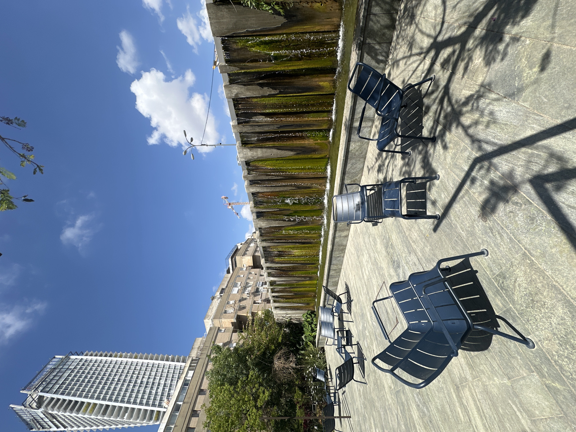 An urban outdoor scene in Tel Aviv, Israel, is depicted on a bright, sunny day. The foreground and midground feature a paved plaza area furnished with several dark blue metal chairs, designed with slatted backs and seats. Strong, elongated shadows of unseen trees and the chairs themselves are cast across the light-colored paving, indicating direct, intense sunlight.
Running parallel to the seating area is a multi-tiered concrete water feature or fountain. Water cascades down its levels, which are notably covered in significant growth of dark green algae and moss, lending a weathered, natural appearance to the structure.
In the background, a mix of city architecture rises against a vivid blue sky. To the left, a tall, modern high-rise building with a distinctive white, grid-patterned facade stands out. Adjacent to it and stretching along the midground are older, light-colored apartment buildings, characterized by numerous external air conditioning units and balconies. Lush green trees and foliage are interspersed among the buildings and alongside the water feature.
The sky above is clear and vibrant blue, dotted with scattered white, fluffy clouds. A lamppost with multiple light fixtures is visible against the sky near the water feature, and a distant construction crane can be glimpsed above the buildings. No people are present, and no visible text is discernible in the frame. The overall atmosphere is peaceful and bright.